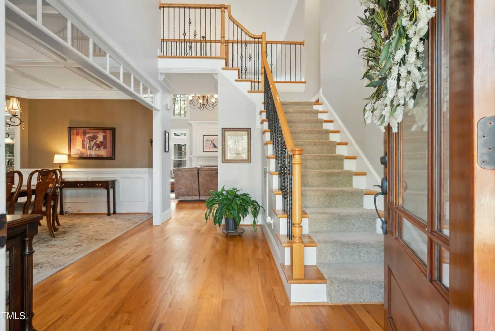A spacious home entryway with hardwood floors, a carpeted staircase with wooden handrails, a potted plant at the base, and decorative wall art. Adjacent rooms are visible, featuring dining and living areas.