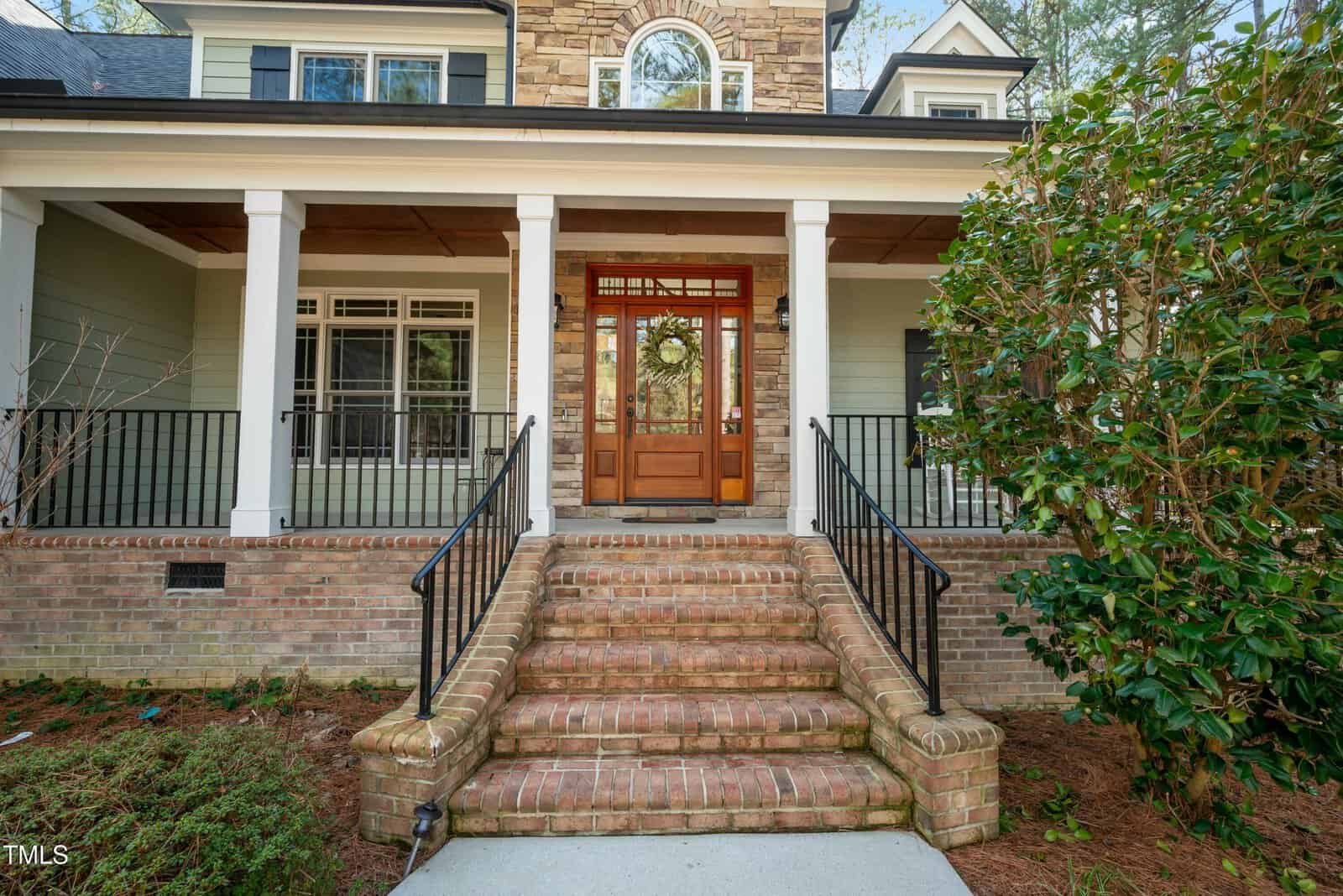 A set of brick steps with black railings leads to a wooden front door with glass panels, surrounded by brick and stone detailing on a house with light green siding and white trim.