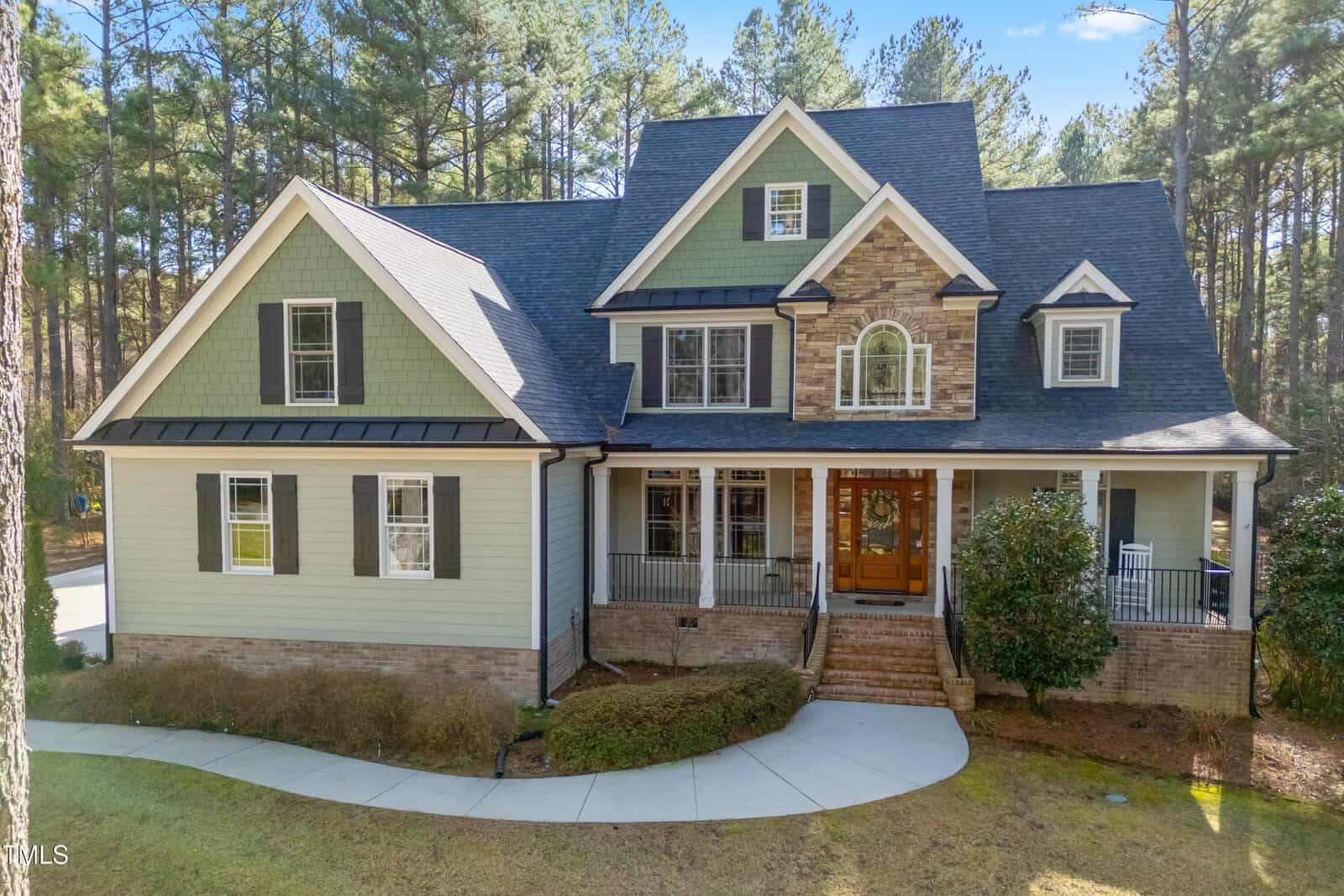 A two-story house with green and beige siding, stone accents, a dark roof, large windows, a covered front porch with white railings, and a brick staircase, set in a wooded area with a curved sidewalk.
