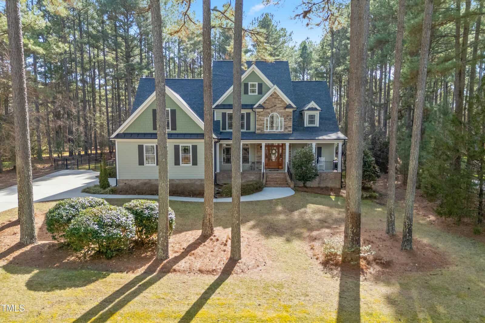 A two-story house with a front porch, gabled roof, and stone accents sits among tall pine trees. There is a curved driveway and landscaped bushes in the front yard, with pine needles covering the ground.