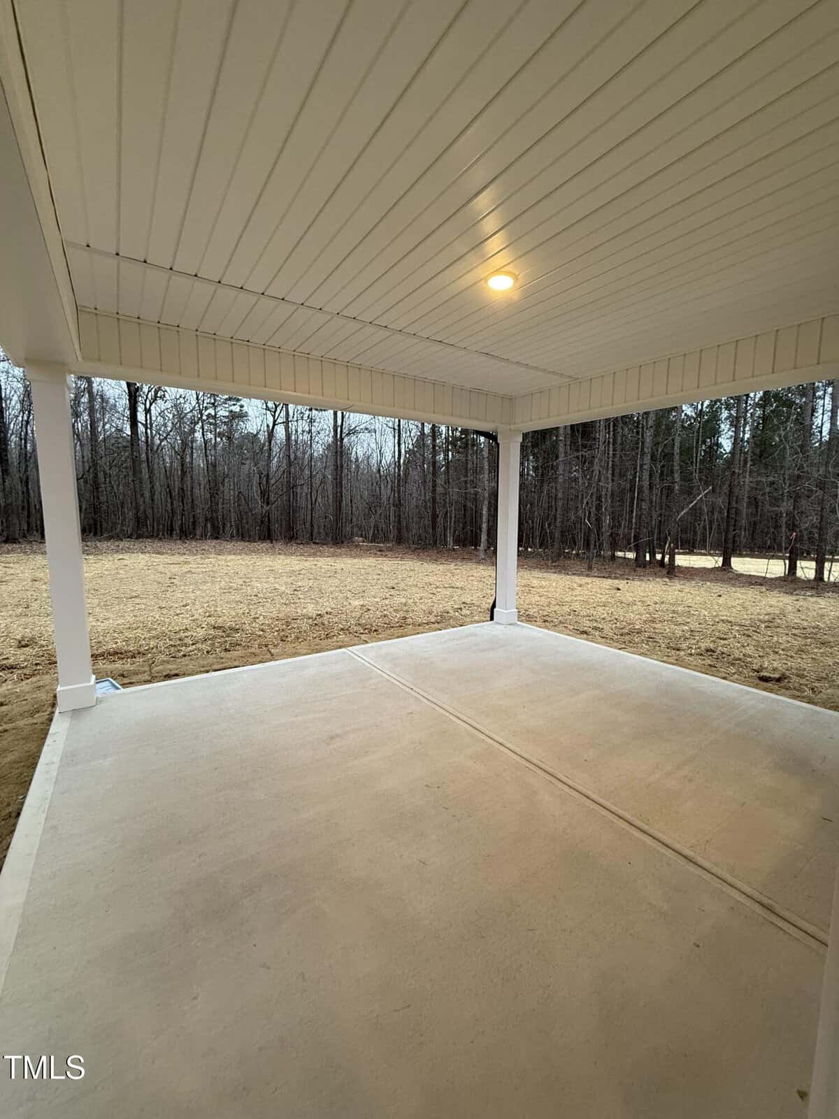 Covered concrete patio with white ceiling, single recessed light, and white support columns. The patio overlooks a grassy yard with bare trees in the background on a cloudy day.