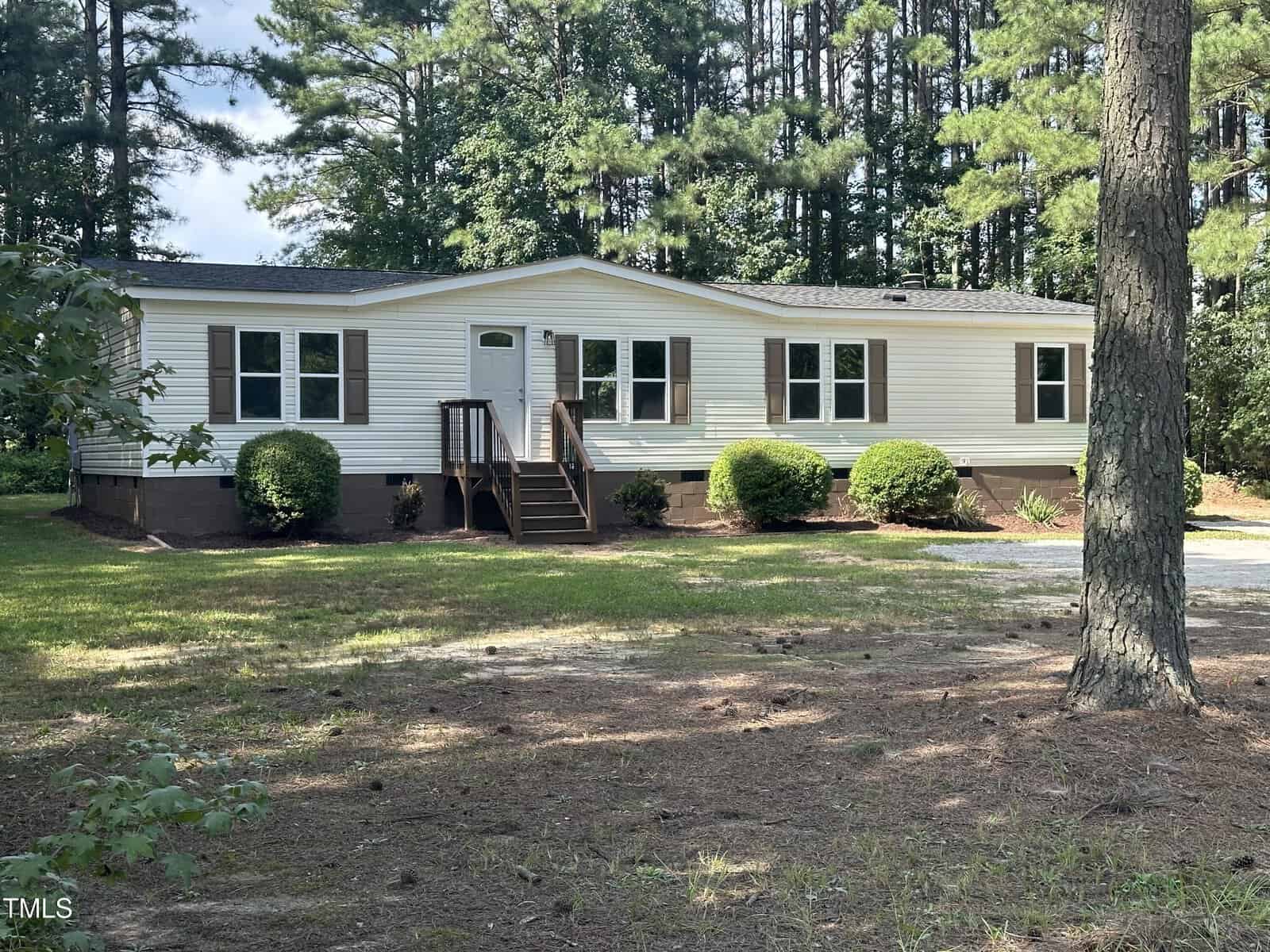 Nestled at 552 Sombrero Lane in Henderson, a white mobile home with a black roof is enveloped by lush greenery and trees. A small wooden staircase leads to the entrance, flanked by bushes. In the foreground, a dirt pathway adds rustic charm to this serene setting.