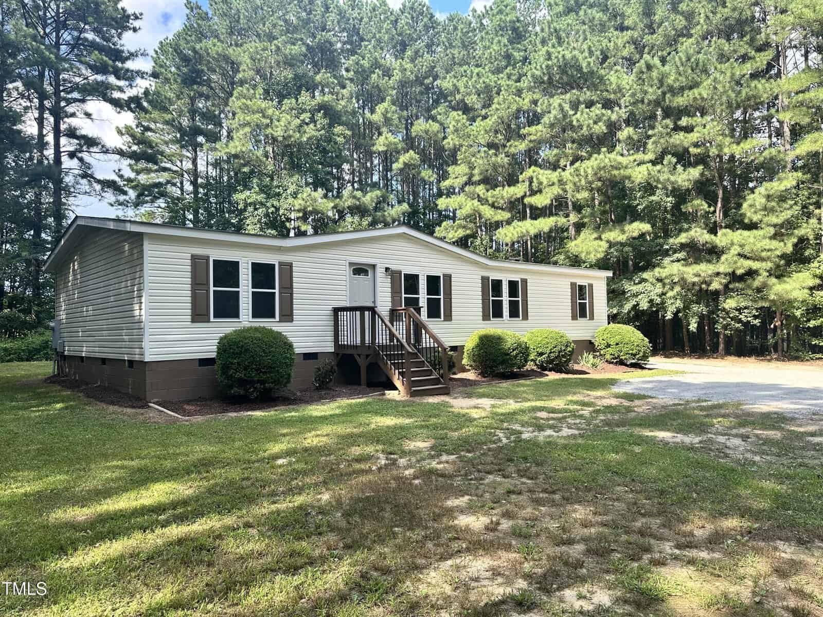 A single-story manufactured home with beige siding and a small wooden porch. It is surrounded by a lawn, bushes, and tall pine trees in the background. A gravel driveway leads to the front of the house. The sky is partly cloudy.