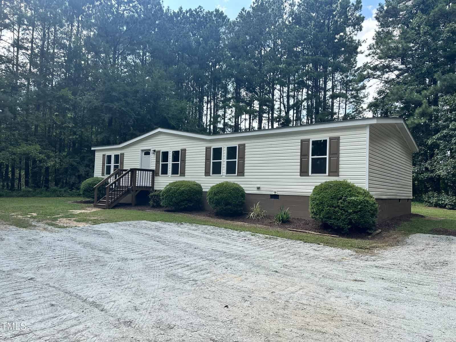 A single-story modular home with light-colored siding and dark shutters is situated on a gravel driveway. The house features a small porch with steps and is surrounded by trimmed bushes and dense trees in the background.