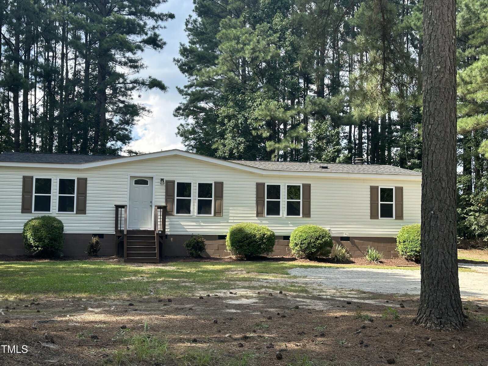 A single-story white manufactured home with brown shutters and a front porch. It is set in front of a wooded area with tall trees. The yard is partially gravel with some grass and shrubs near the house.