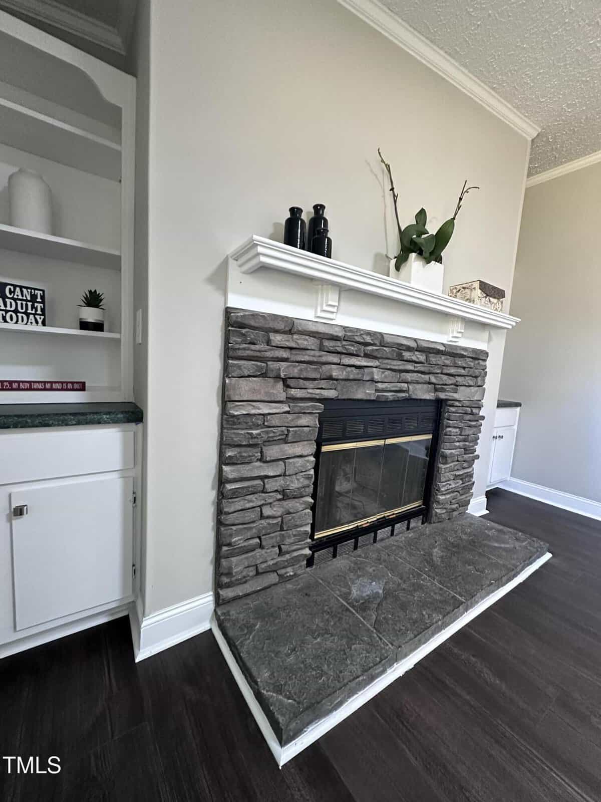 Stone fireplace with a white mantel, decorated with a plant and decorative items. Shelves to the left hold books and a sign reading "Can't Adult Today." Dark wooden floor and light gray walls complete the room's decor.