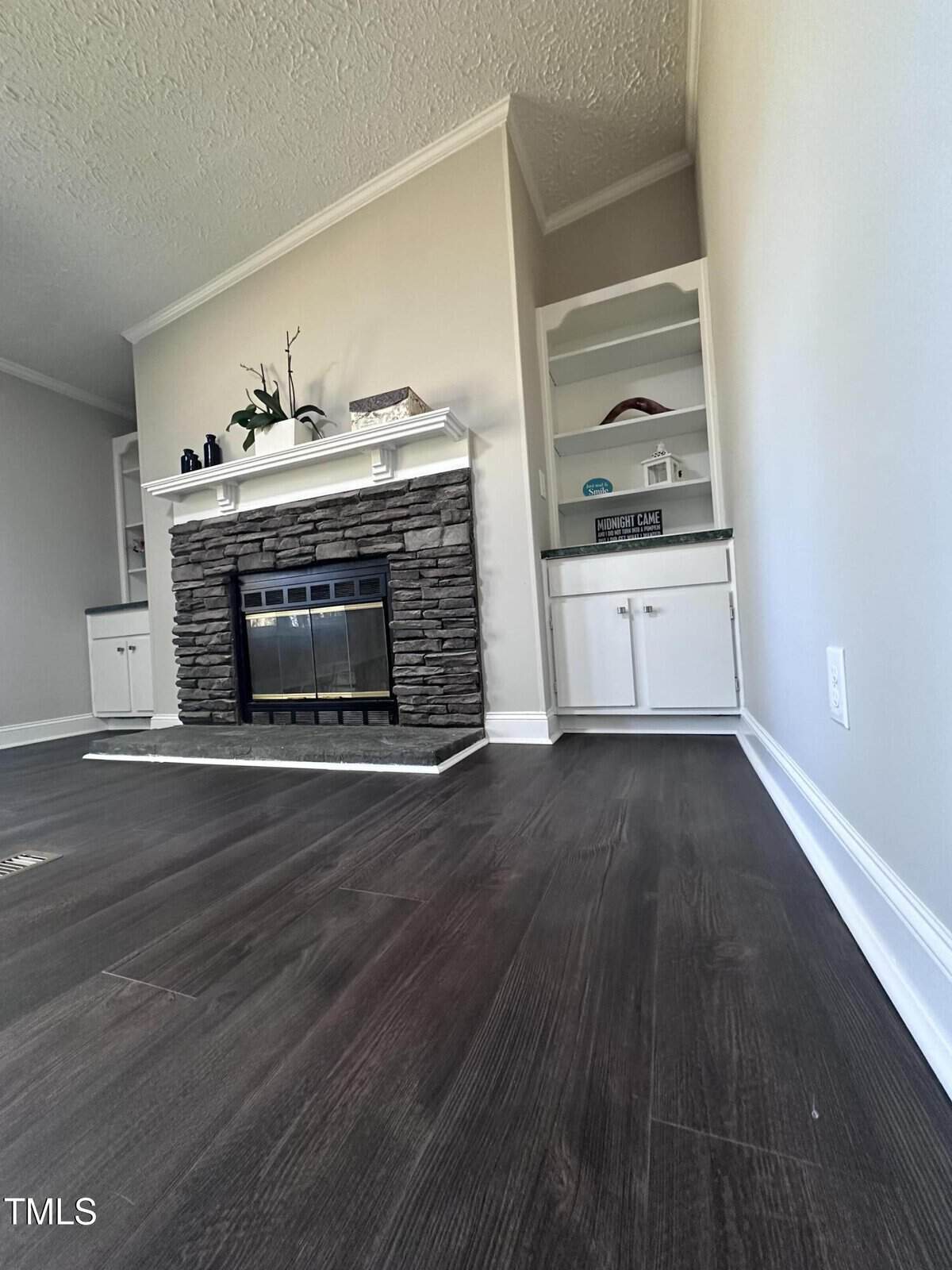 Living room with dark hardwood flooring, featuring a stone fireplace with a black insert. Above the mantel are decorative items. Built-in white shelves are located to the right of the fireplace. Walls are painted light gray.