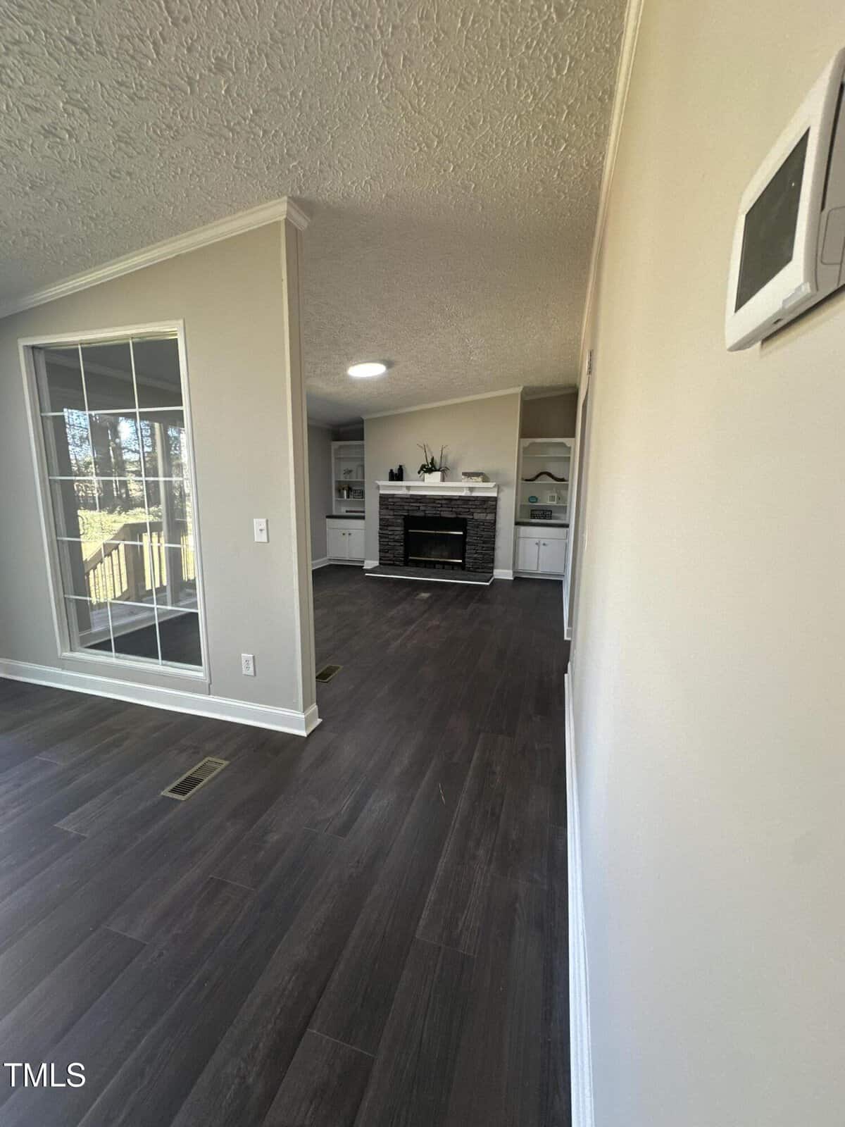 View of a room with dark wood flooring and light-colored walls. A window with multiple panes is on the left, and a fireplace with a stone surround is visible ahead. Built-in shelves flank the fireplace, and a thermostat is mounted on the right wall.