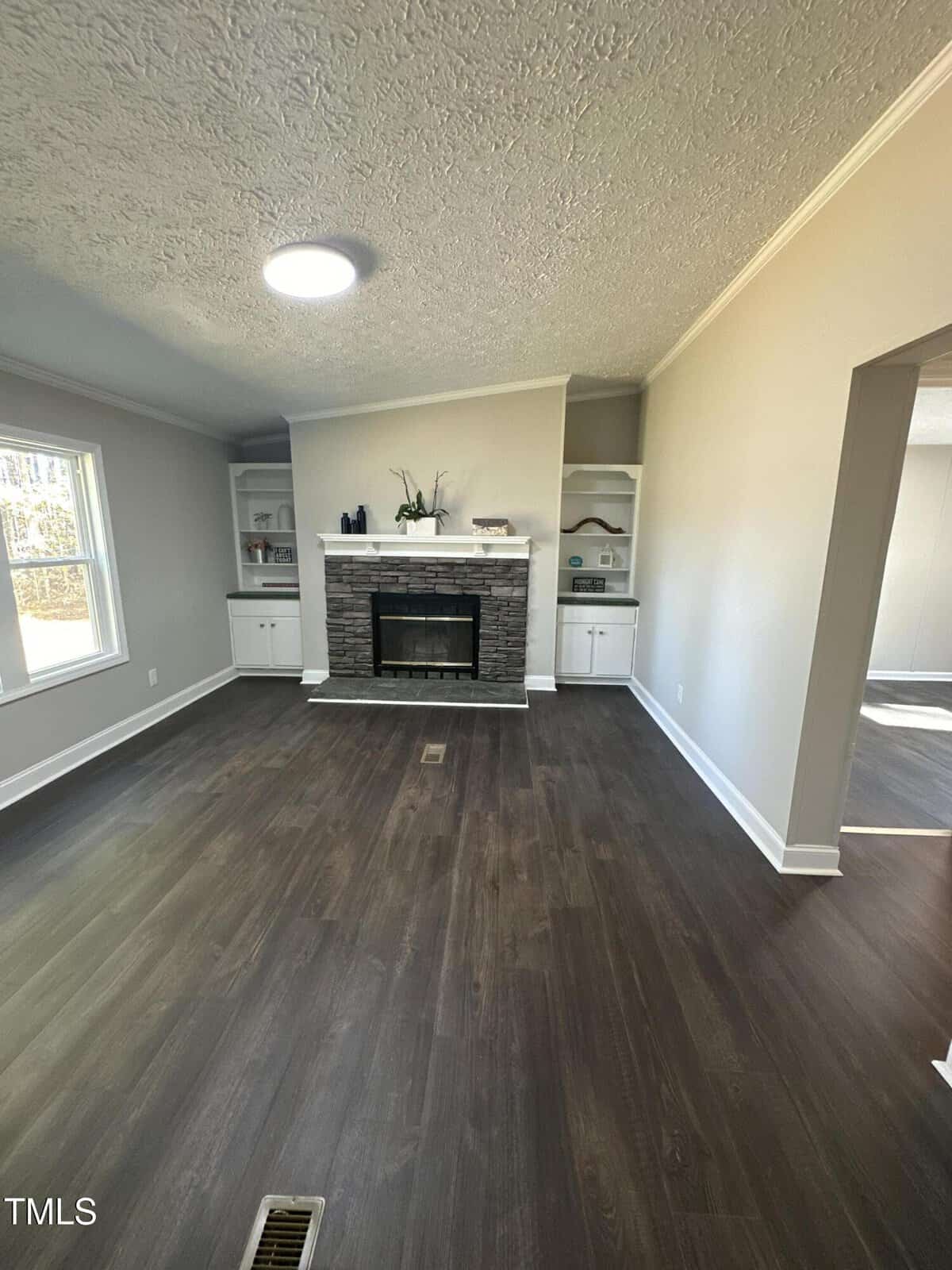 A living room with dark wood flooring, featuring a stone fireplace centered on the far wall. There are built-in shelves on either side of the fireplace. Natural light enters from a window on the left, highlighting the light-colored walls.