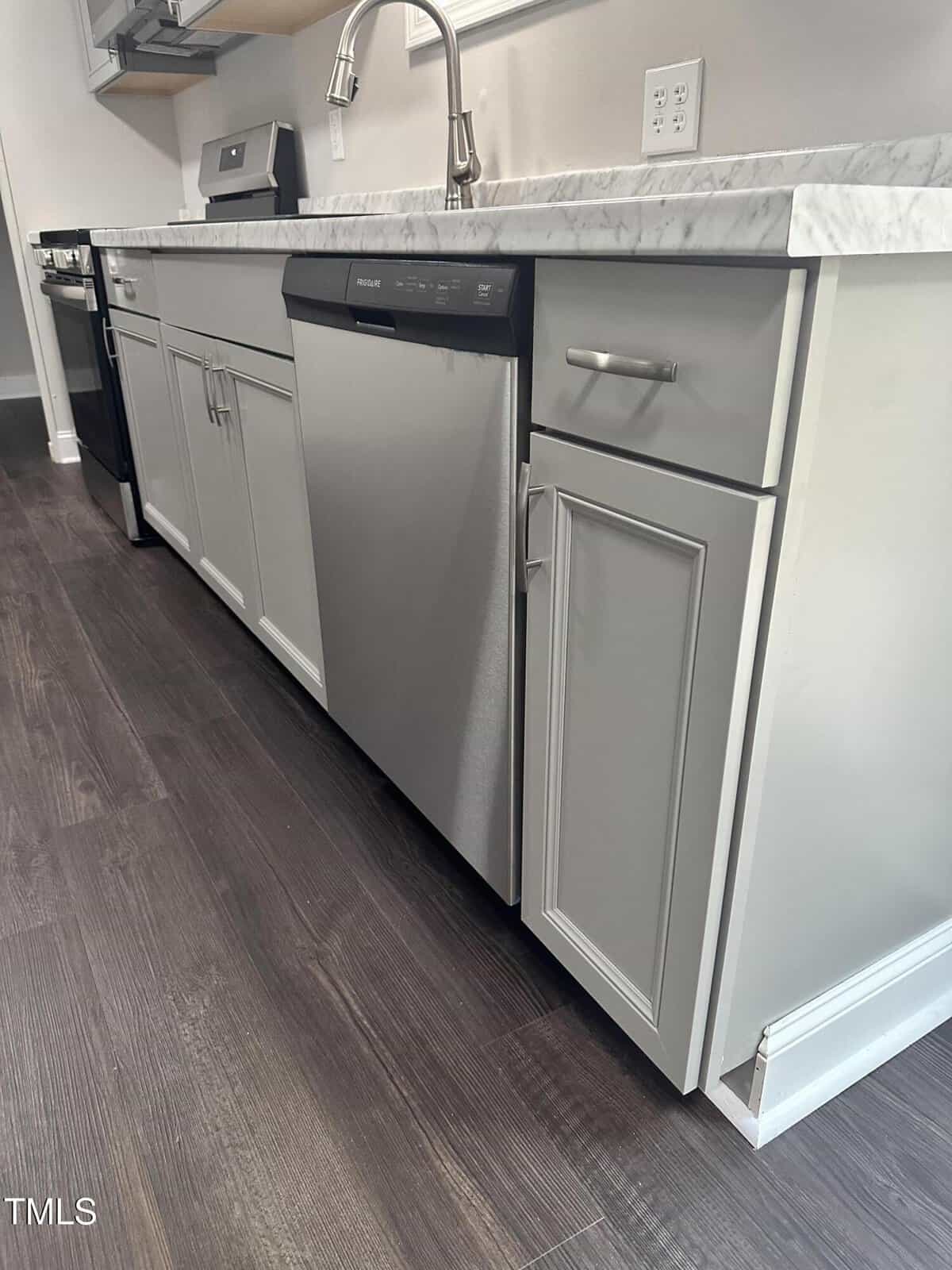 A kitchen with a marble countertop, featuring a stainless steel dishwasher, oven, and sink. The cabinets are grey, and the floor is made of dark wood. The setting is clean and modern.