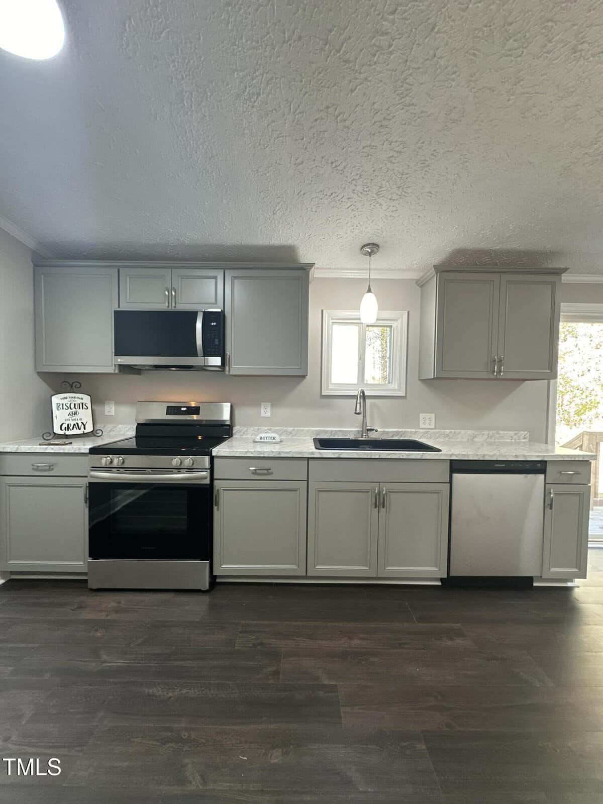 Contemporary kitchen with grey cabinets, white marble countertops, and stainless steel appliances, including a microwave, oven, and dishwasher. A light hangs above the sink, and a window offers natural light. Dark wood flooring is visible.