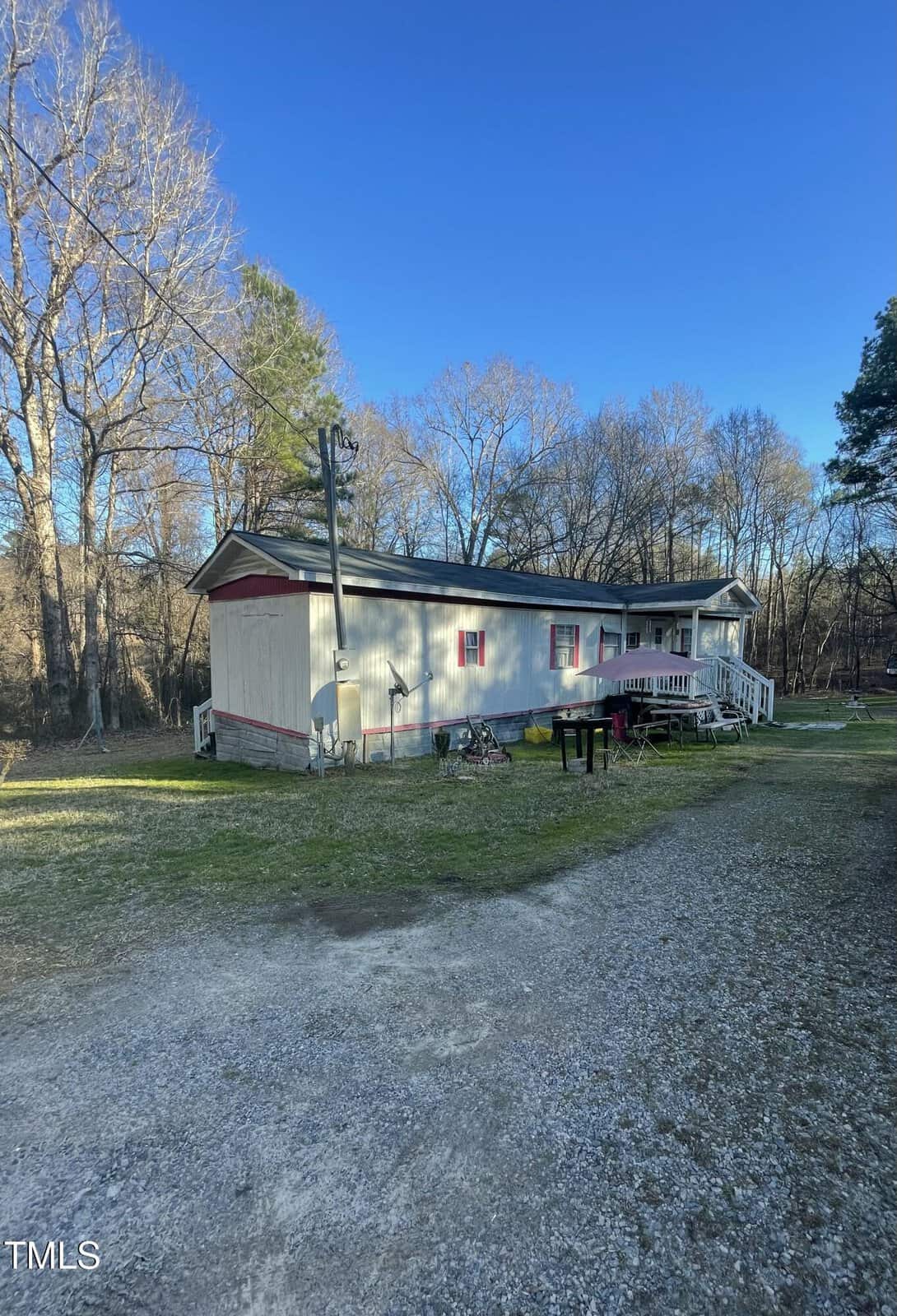 A single-story light gray mobile home with pink window frames is set on a grassy lot with a gravel driveway and surrounded by leafless trees under a clear blue sky.