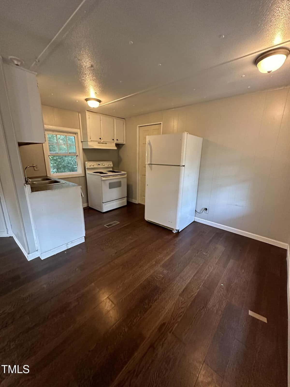 A small kitchen with white cabinets, a white stove, a white refrigerator, a stainless steel sink, and dark wood floors. There are beige walls, a window, and ceiling lights. The space is mostly empty.