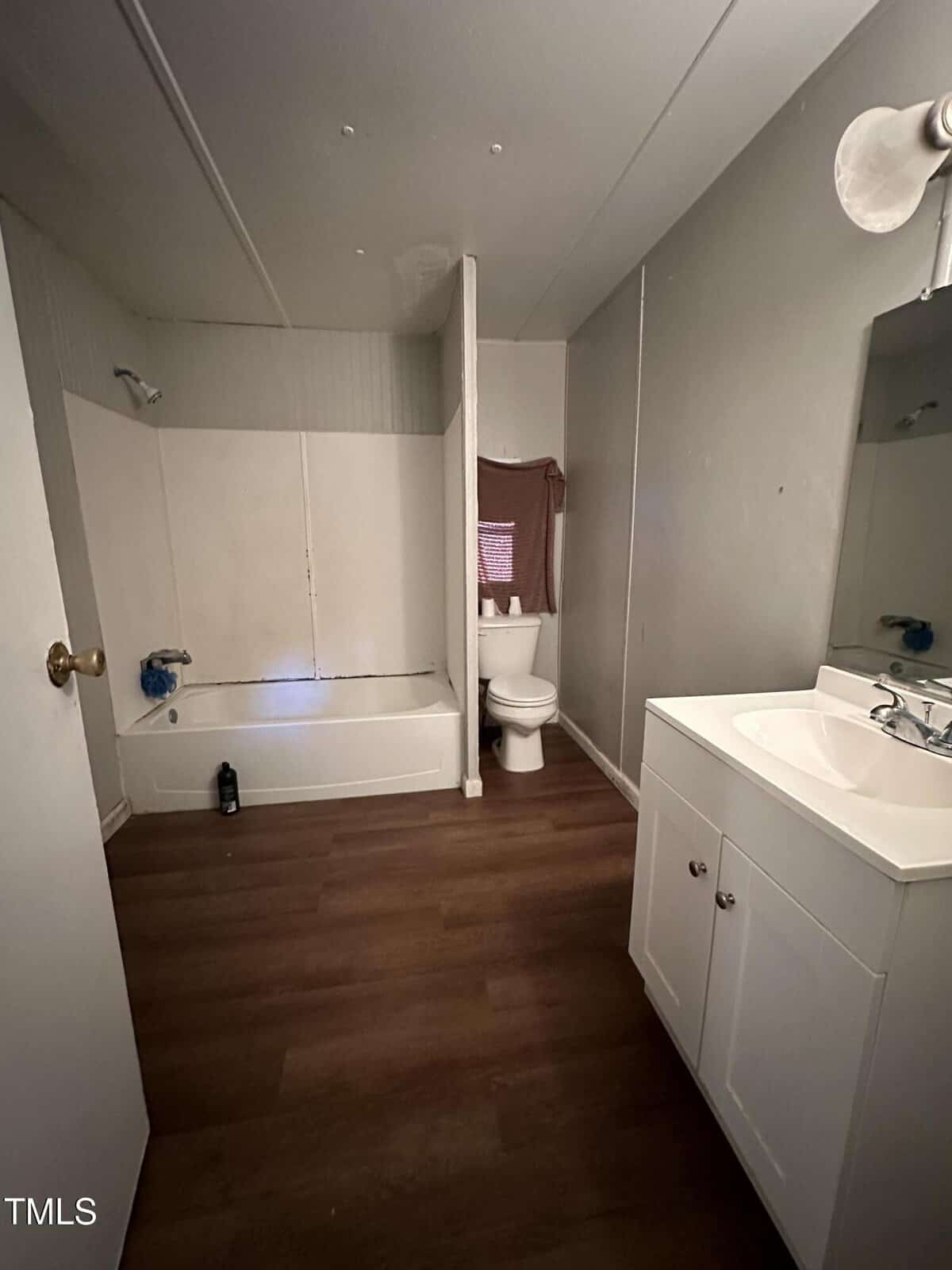 A bathroom with a white bathtub and shower on the left, a toilet next to a window with a curtain in the center, and a white sink with a mirror and light fixture on the right. The floor is dark wood.