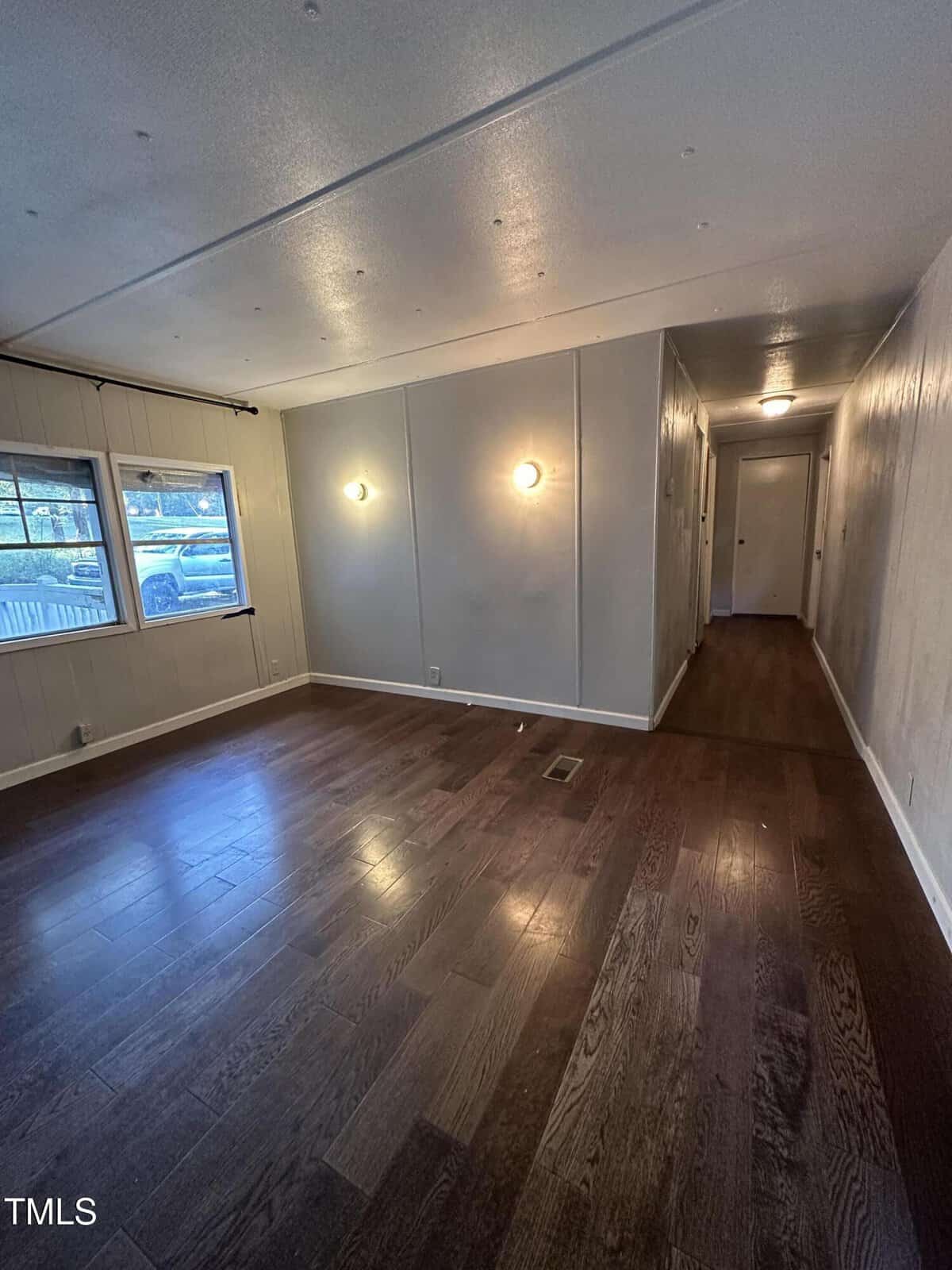A narrow living room with dark wood floors, light gray walls, two round wall lights, a window on the left showing a parked car outside, and a hallway leading to a closed door in the back.