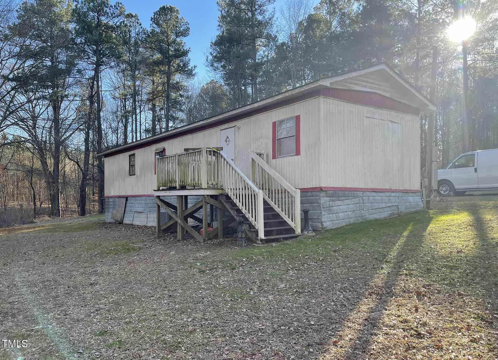 A beige, single-wide mobile home with red trim sits on stacked blocks. Wooden stairs lead to the front door. A white van is parked nearby. There are trees in the background and sunlight streams from the right.