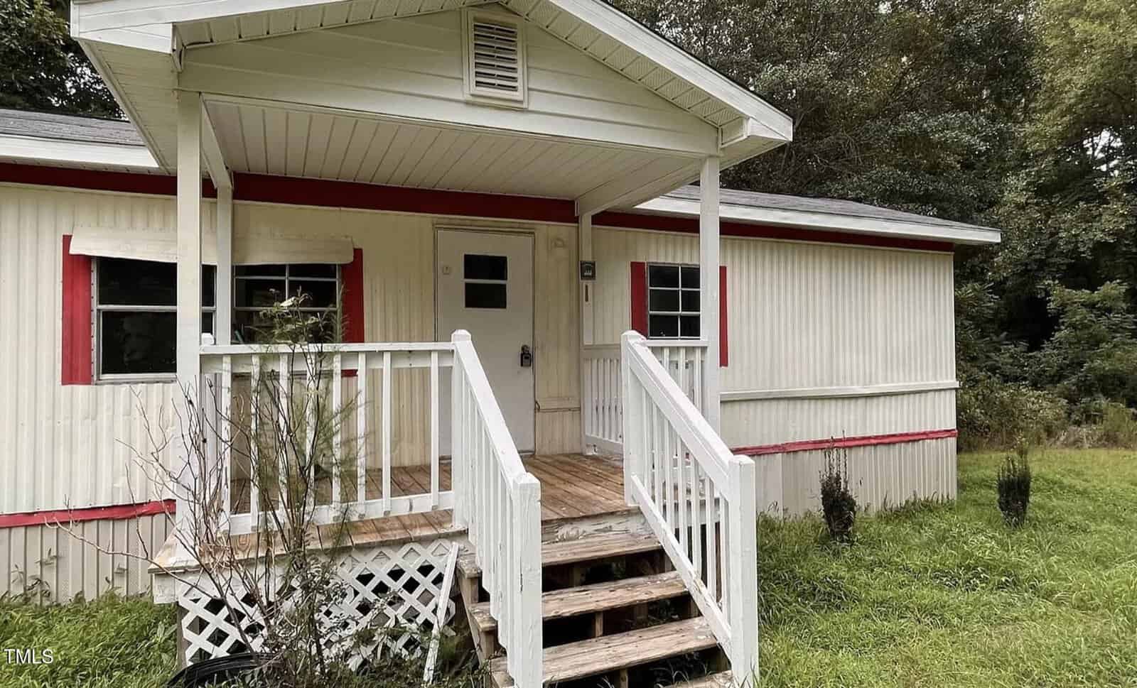 A small, single-story house with white siding and red trim, featuring a covered porch with white railings and steps, overgrown grass, and a few small shrubs in the front yard. Trees are visible in the background.