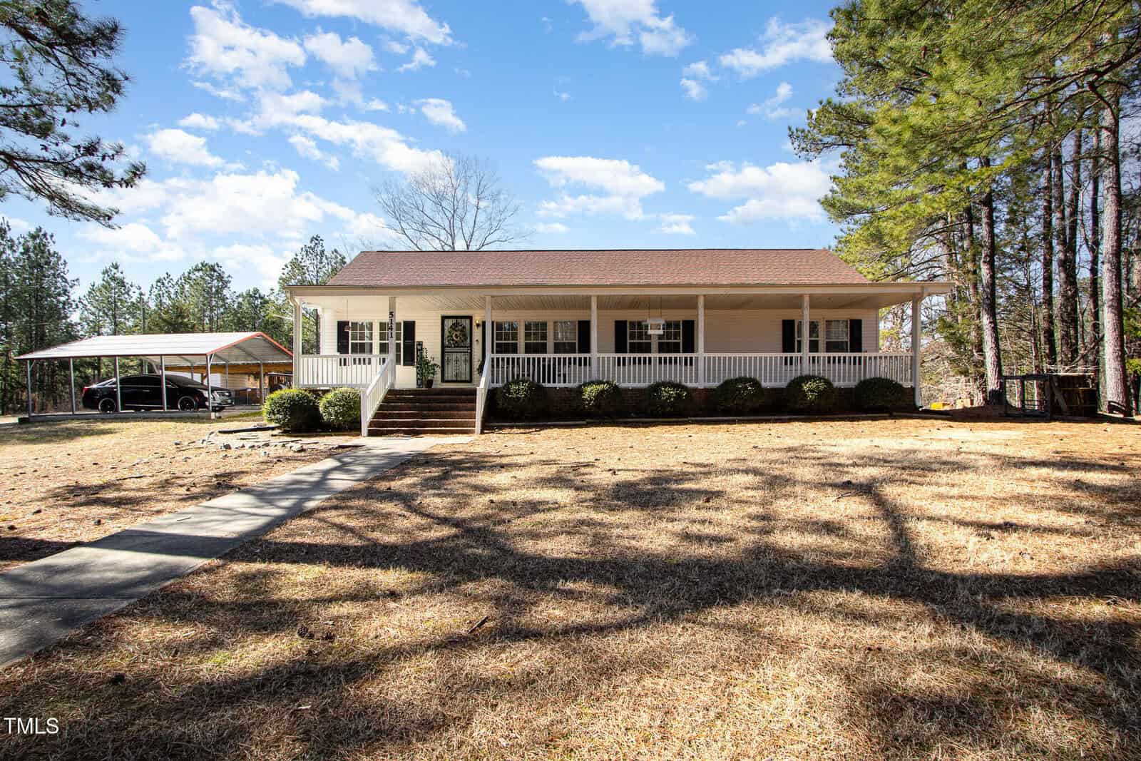 Nestled along 5141 NC-39 Highway Highway in Henderson, this charming single-story house features a large front porch with white railing, surrounded by trees. A carport sits to the left, and a concrete path leads to the steps. The grassy yard enjoys dappled shadows under the partly cloudy sky.