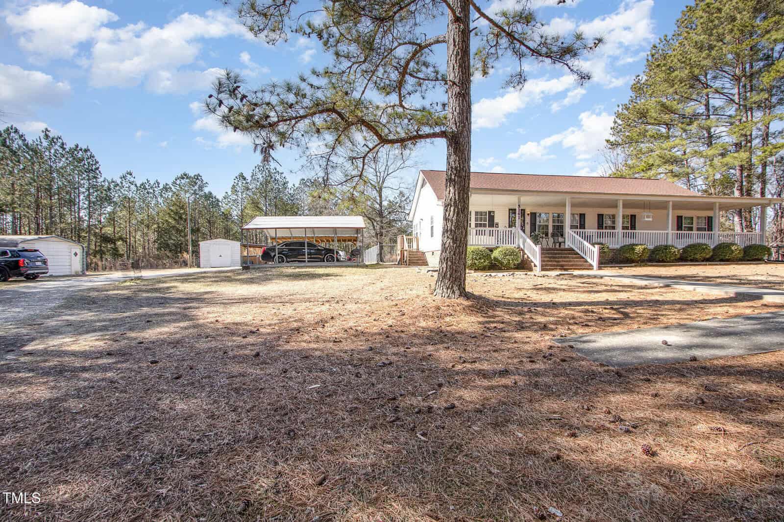 A single-story house with a covered porch is surrounded by a spacious yard with pine trees. A boat is parked under a carport to the left, and a shed is visible nearby. The sky is partly cloudy.