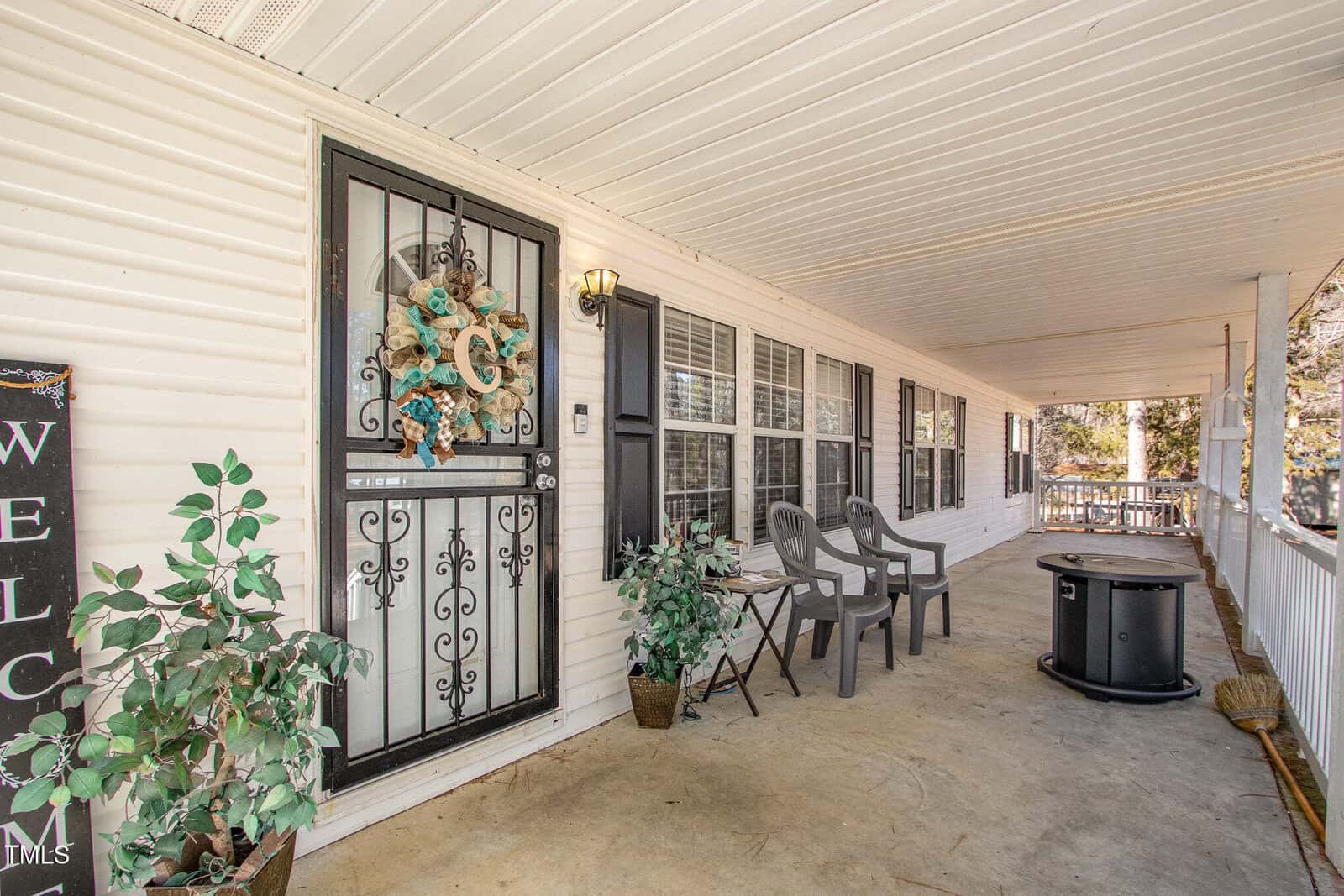 A covered porch with a metal door featuring a decorative wreath and a letter "C." Three plastic chairs and a small table are on the porch. A welcome sign and potted plants are by the entrance. White railings run along the porch edge.