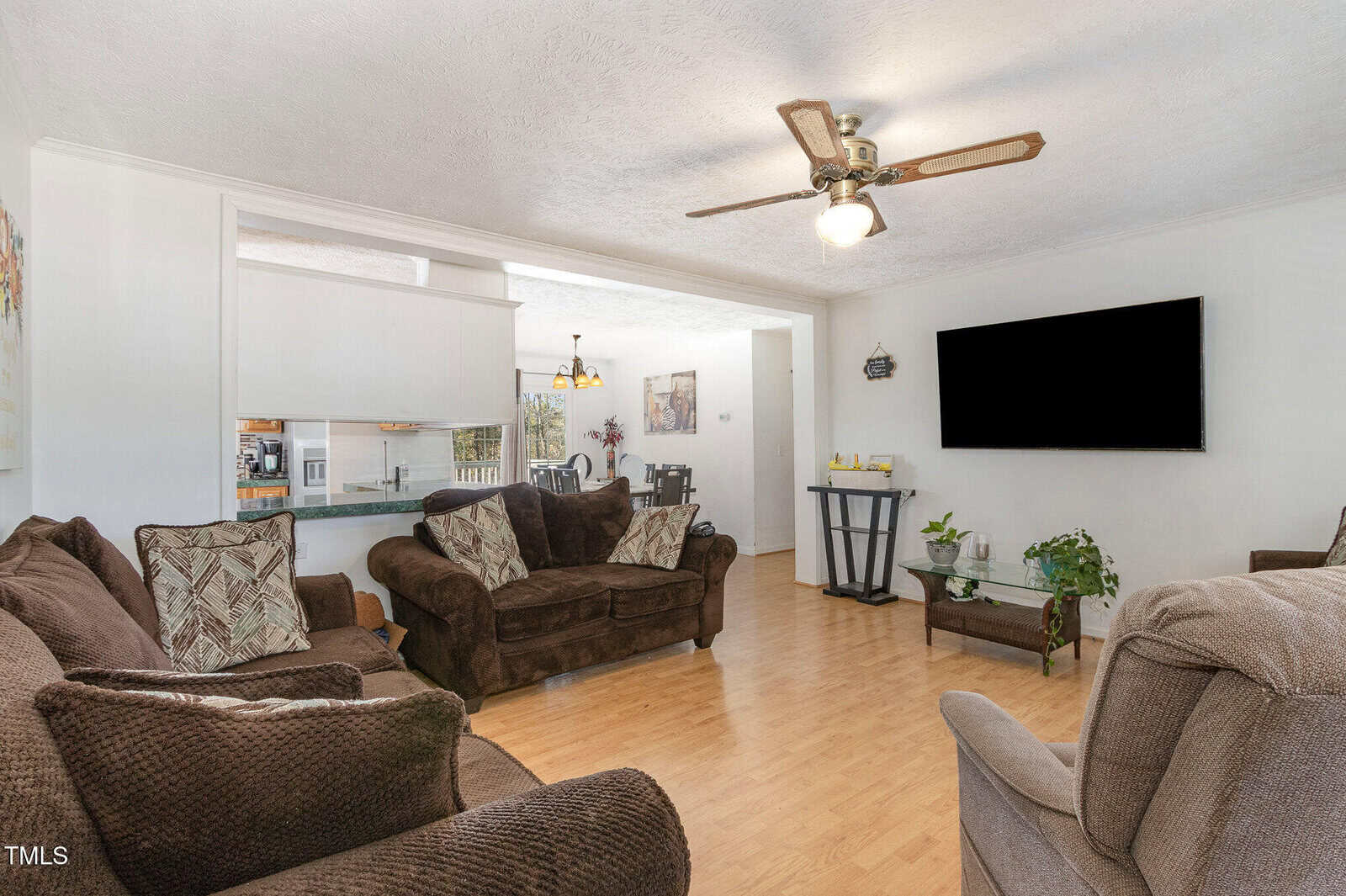 Living room with wooden floor, brown sofas, and a wall-mounted flat-screen TV. A ceiling fan with a light fixture is above, and several plants and decorative items are on a stand. A dining area is visible in the background.