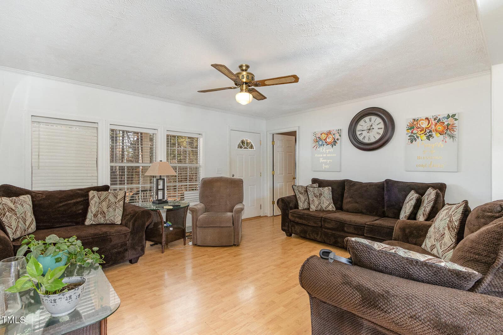 A living room with brown sofas, patterned cushions, and a brown recliner. A clock hangs on the white wall, flanked by floral artwork. The wooden floor contrasts with the white walls. A ceiling fan with a light is centered above.