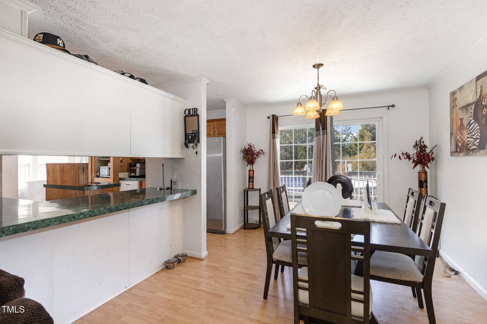 A dining room with a rectangular table and six chairs on a wooden floor. A light fixture hangs above the table. To the left, a kitchen area is visible with a breakfast bar and green countertop. A large window at the back lets in natural light.