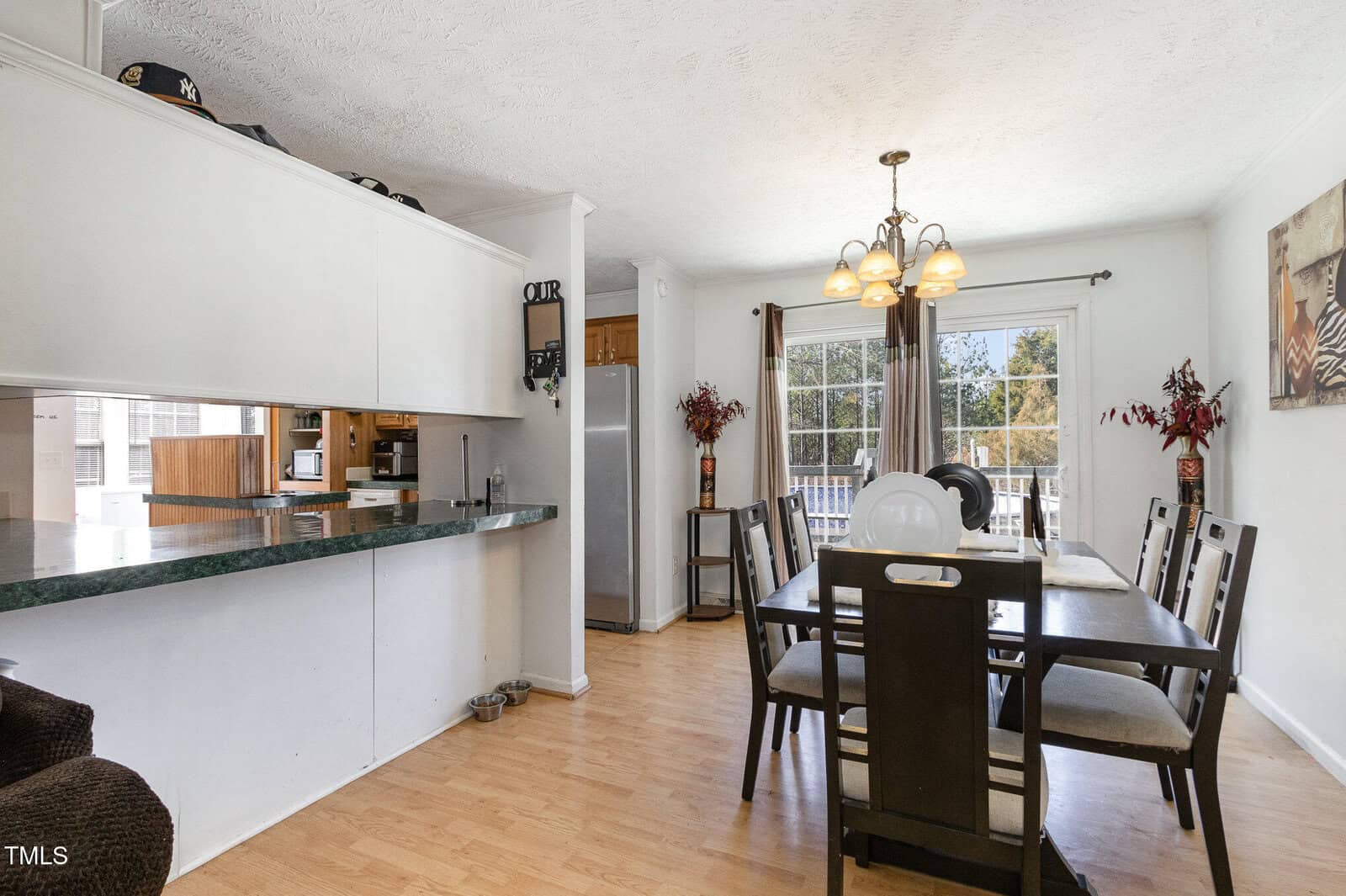 A dining room features a dark wood table with six chairs, a hanging light fixture, and a window with curtains. The open kitchen has a counter, stainless steel refrigerator, and wooden cabinets. Decorative items are placed around the room.
