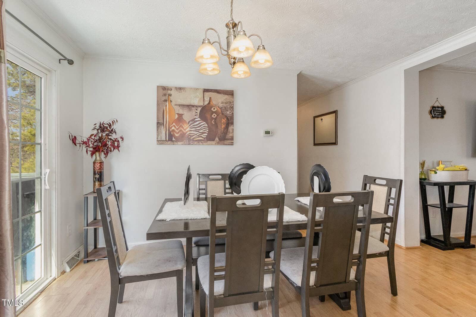 Dining room with a rectangular table set for six, featuring gray cushioned chairs. A chandelier hangs above the table. Large painting on the wall, and sliding glass doors to the left. A sideboard with a mirror is visible in the adjacent room.