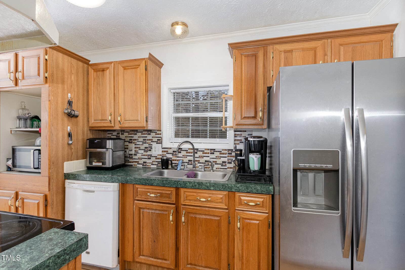 A kitchen with wooden cabinets, a stainless steel refrigerator, a dishwasher, a coffee maker, and a microwave. The sink is under a window with a tile backsplash. A small section of the stove is visible on the left.