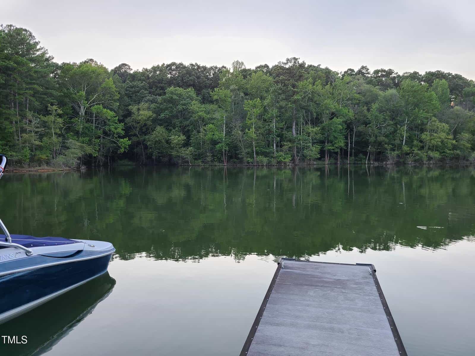 A calm lake is surrounded by dense, leafy trees. A wooden dock extends into the water from the foreground, and a blue boat is moored to the left side of the dock. The reflection of the trees is visible on the lake's surface.