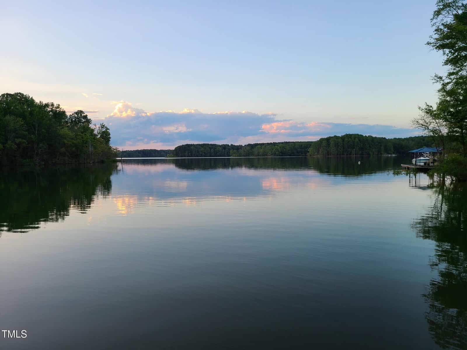 A calm lake reflecting the sky and surrounding trees during sunset. The sky has soft clouds with hints of pink and blue. Trees line the horizon, and a small structure is visible on the right, near the water.