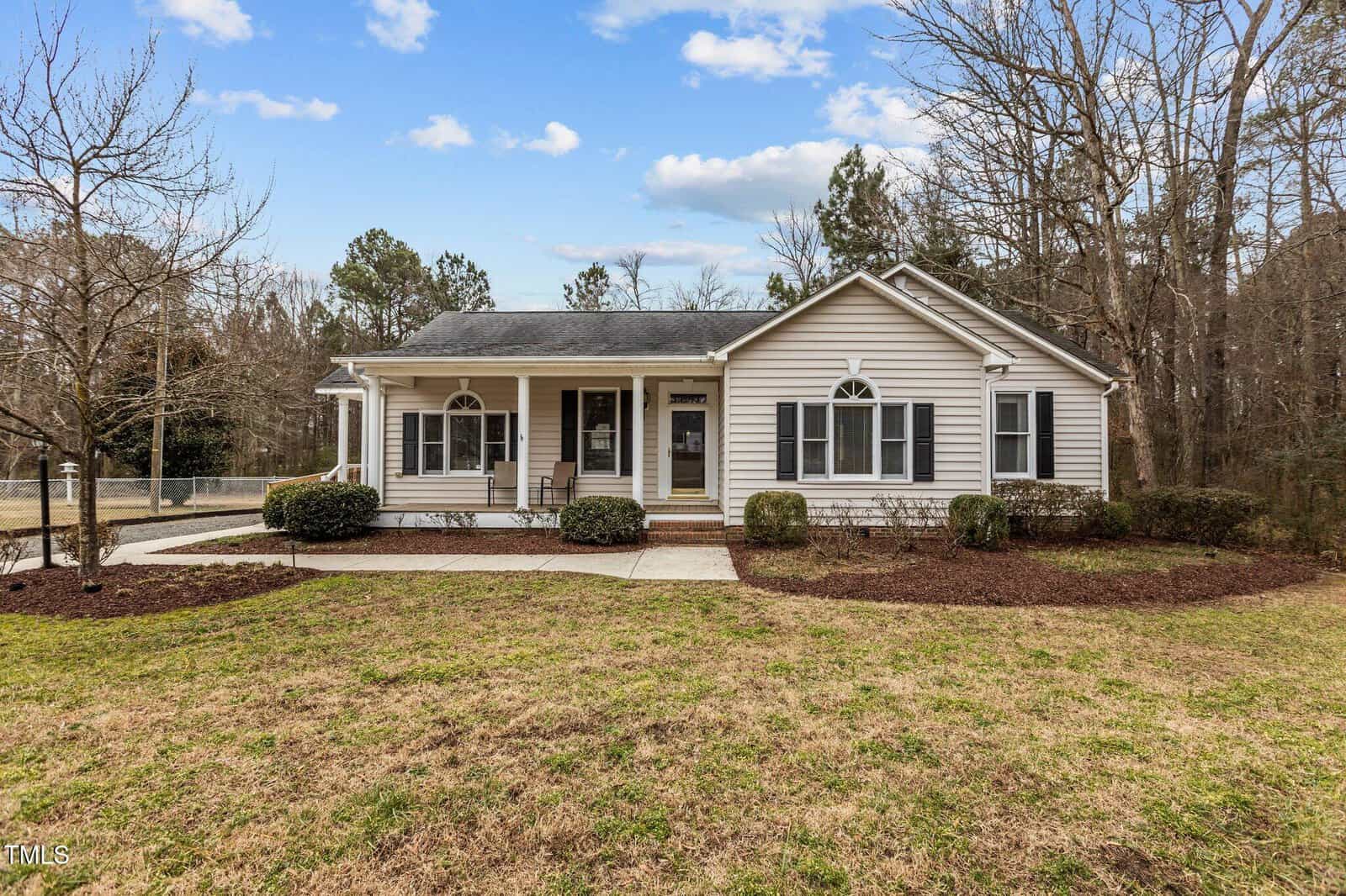 Single-story house with white siding and dark shutters, featuring a wide front porch and multiple windows. Surrounded by a lawn and leafless trees, the house is set against a background of more trees. The sky is partly cloudy.