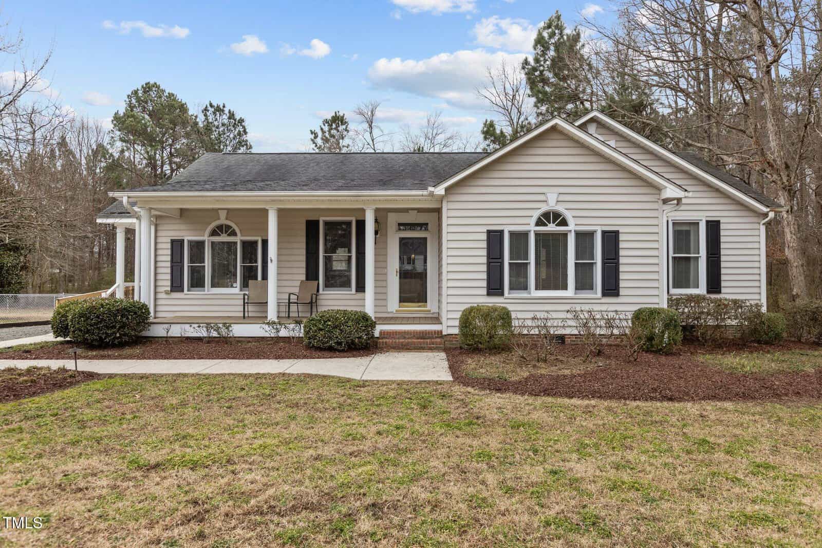 A single-story house with white siding and dark shutters is shown. It has a covered front porch with two chairs. The front yard features a walkway and some shrubs. The background includes trees and a partly cloudy sky.