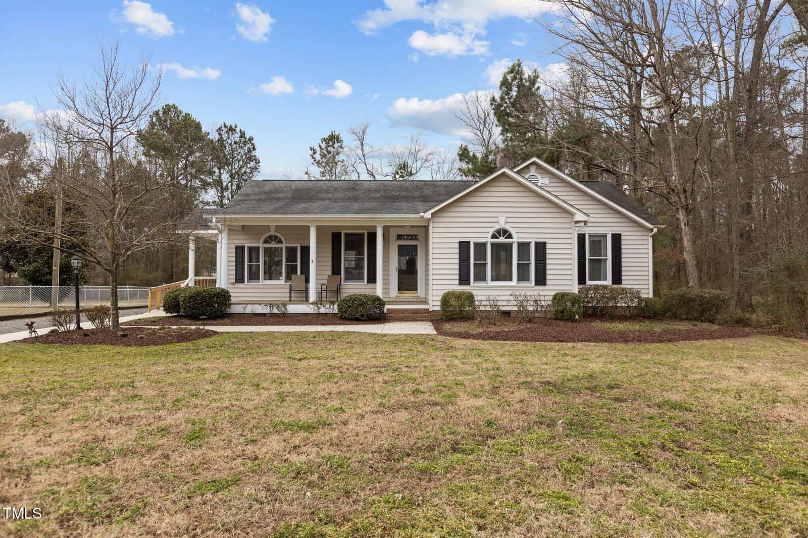 Single-story house with light-colored siding, a front porch, and dark shutters. It is surrounded by a grassy yard and a few leafless trees. The sky is partly cloudy. A driveway is visible on the left side.