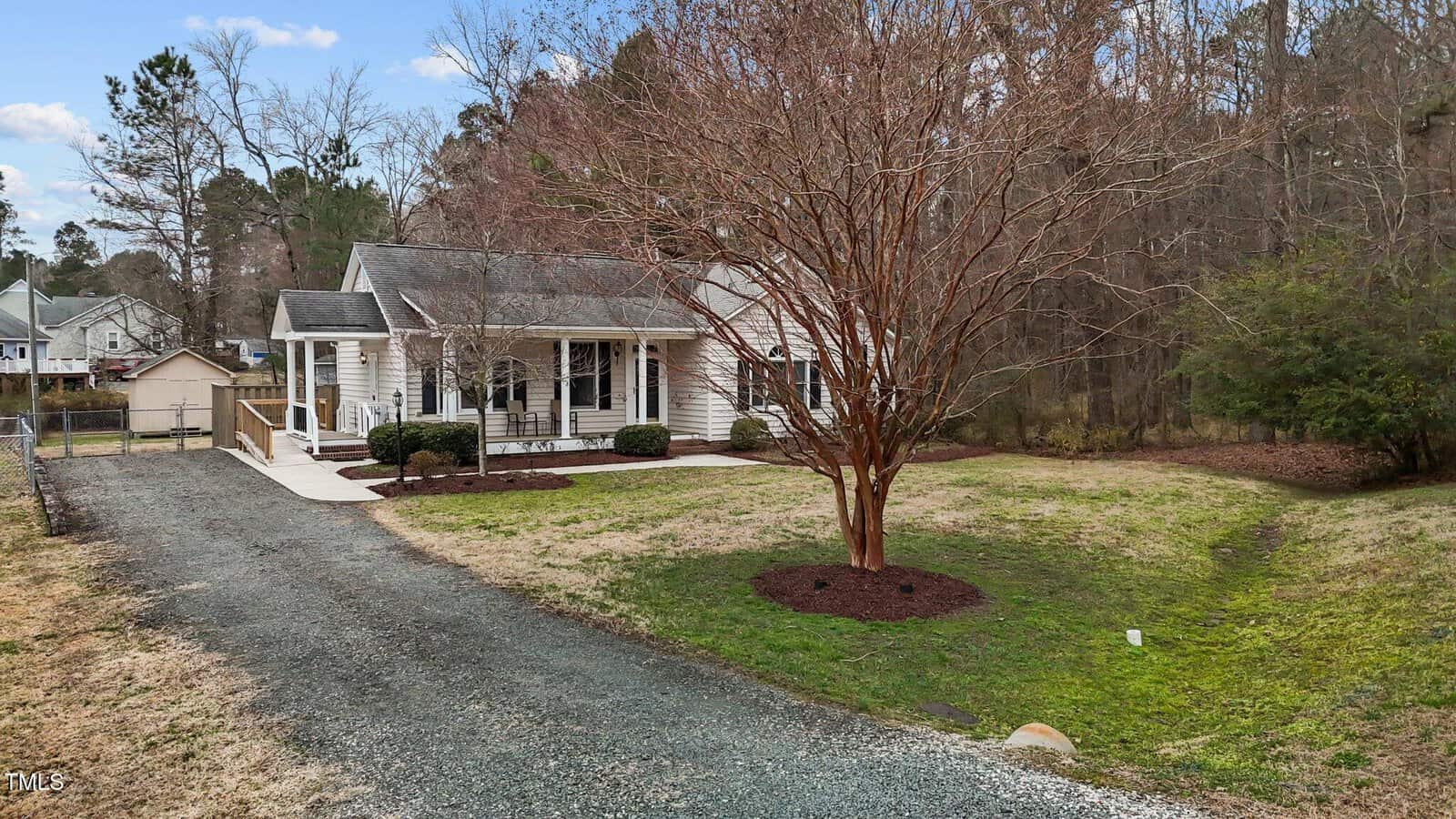 A small, single-story house with a gable roof and a covered front porch sits amid a grassy lawn. A leafless tree is in the front yard, and a gravel driveway leads to the house. Trees and bushes surround the property, with a few neighboring houses visible.