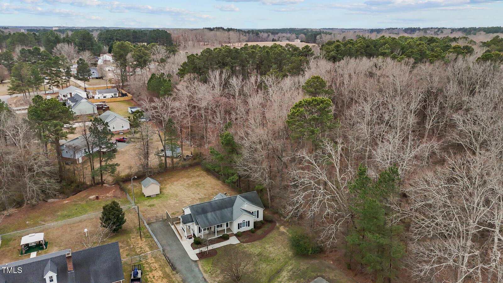 Aerial view of a residential area with a mix of detached houses and wooded areas. The landscape features bare trees, a few pine trees, and grassy yards. The sky is partly cloudy, and there are a few roads running through the neighborhood.