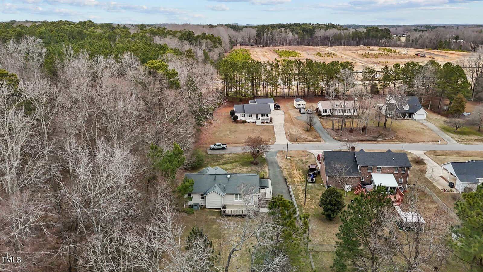 Aerial view of a suburban neighborhood with several houses surrounded by trees. The area includes a mix of leafless and evergreen trees, a few cars parked on driveways, and a cleared field in the distance under a cloudy sky.