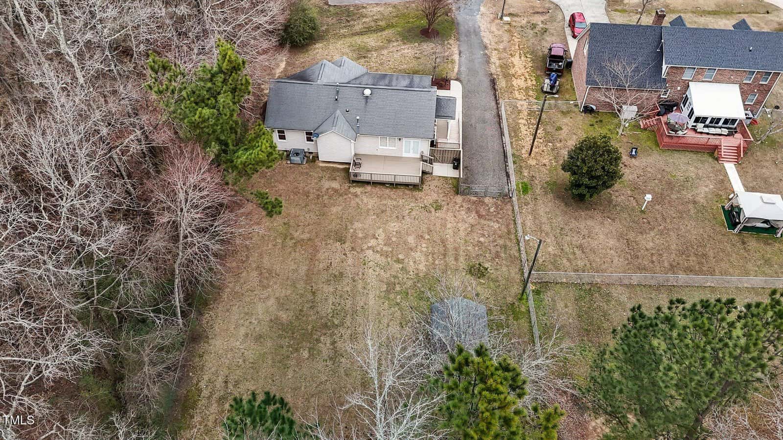 Aerial view of a residential area showing two houses. One house is light-colored with a dark roof, surrounded by trees and an open yard. The other house is brick with a deck. A driveway and small road run between the houses.