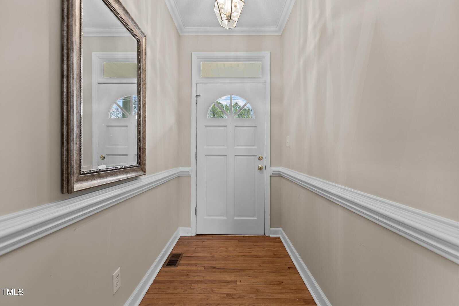 A narrow hallway with beige walls features a white entrance door with a small window. A large framed mirror hangs on the left wall. The floor is wooden, and there is a decorative crown molding along the walls. A pendant light fixture hangs above.