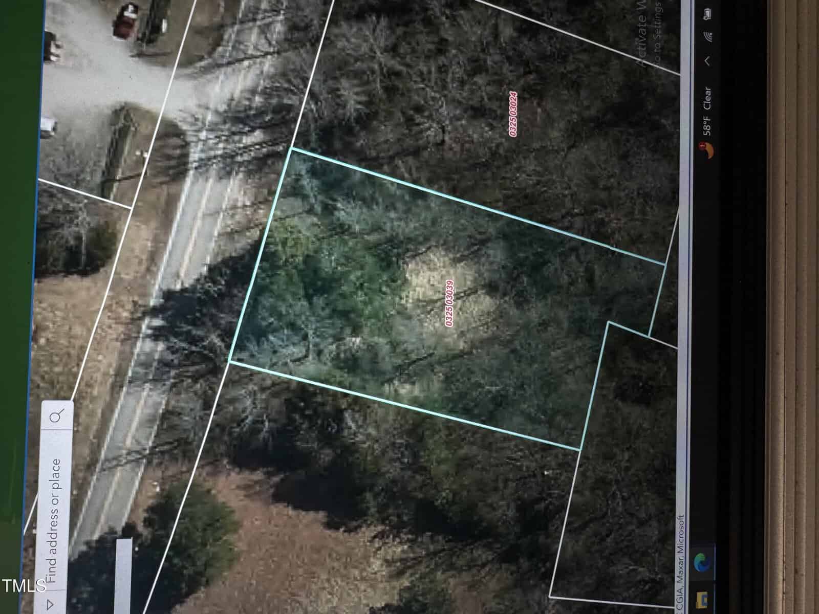 Aerial view of a densely wooded area bordered by thin, light blue lines indicating property boundaries. A road runs along the left side, with additional bordered sections nearby. Some sparse trees are visible outside the main wooded section.
