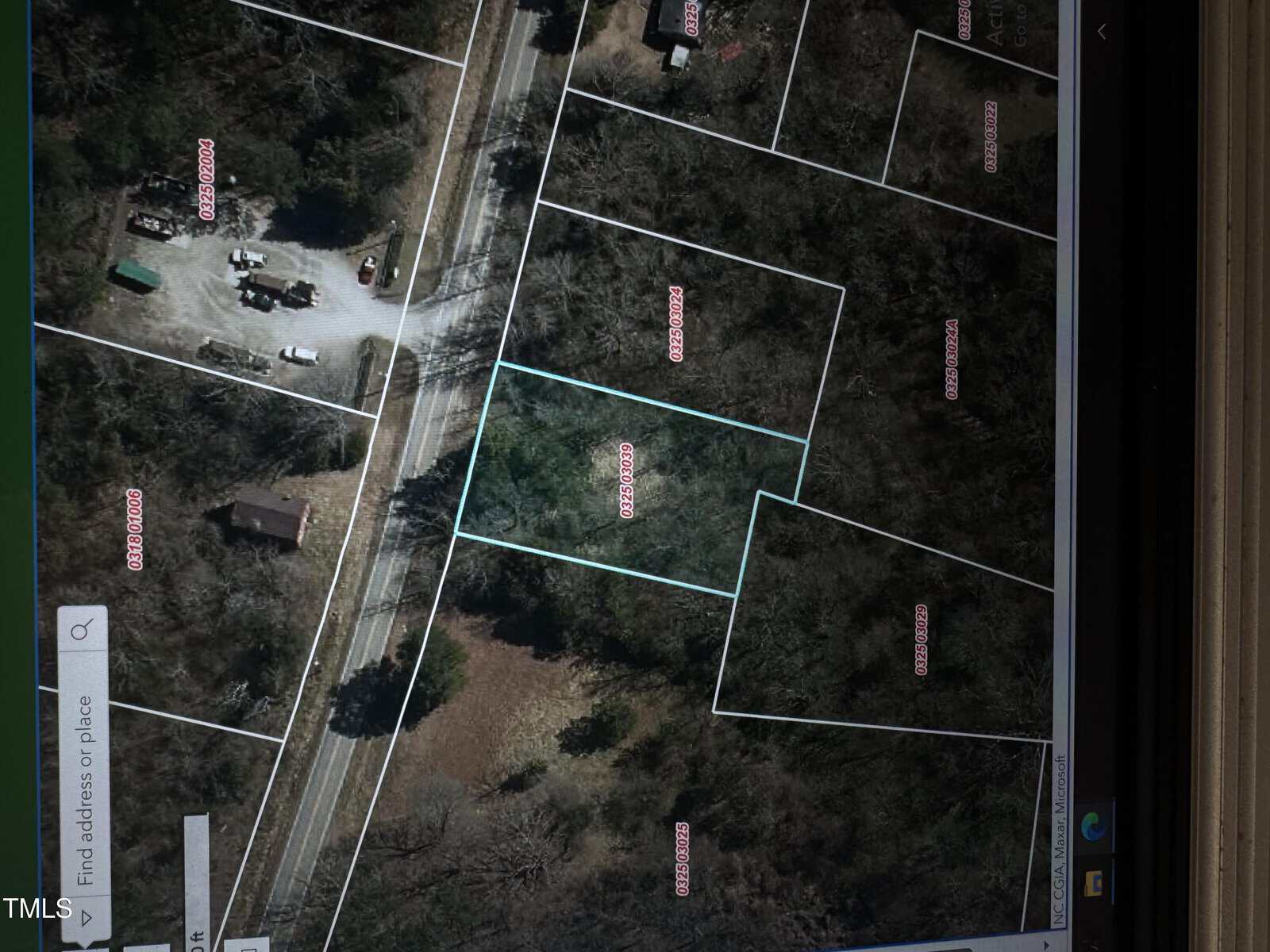 A satellite view of a wooded area with outlined land plots in white. A road runs through the upper section, and various plot numbers are labeled in pink. One plot is highlighted in light blue. Adjacent properties and trees are visible.