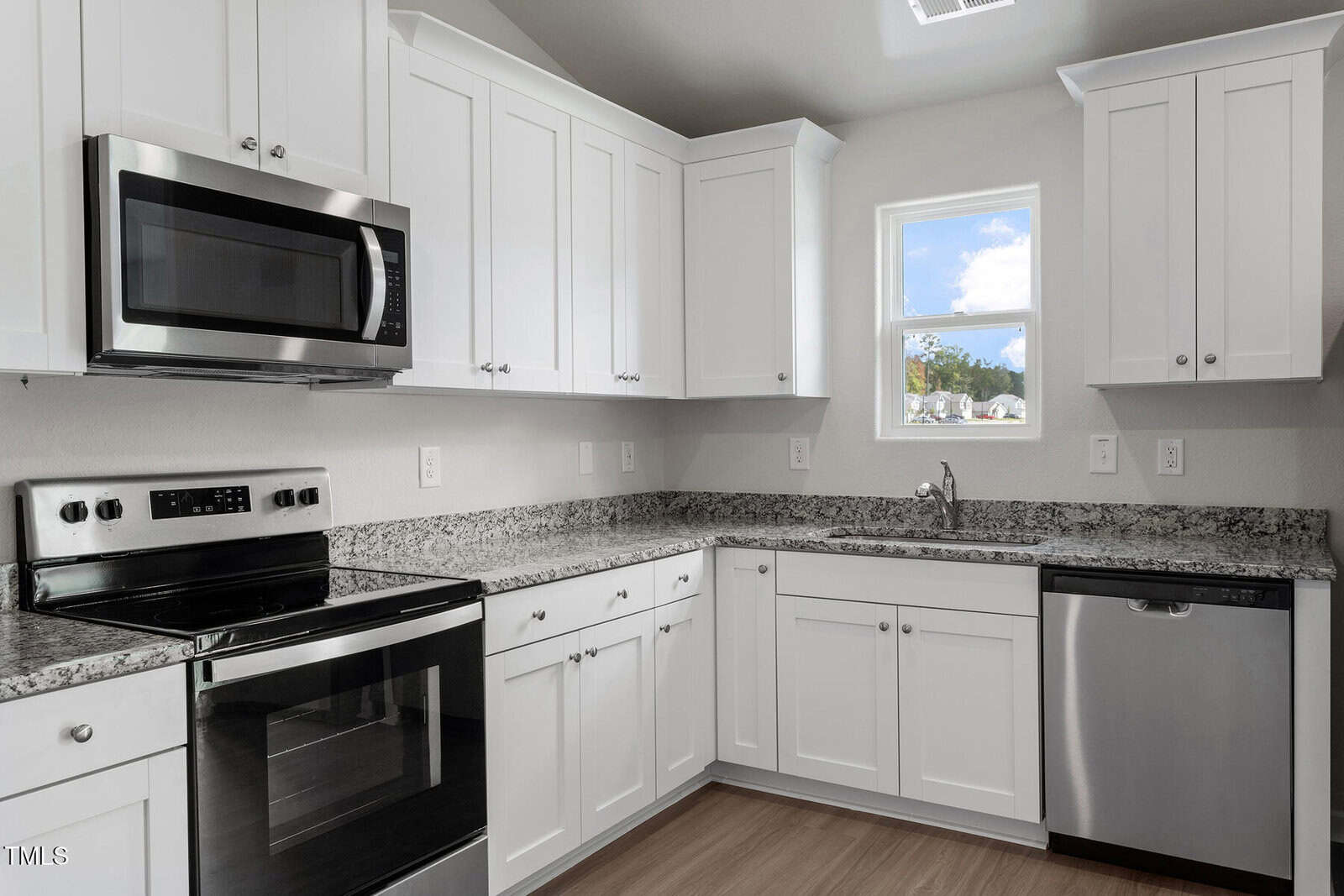 A modern kitchen featuring white cabinets, stainless steel appliances including a microwave, oven, and dishwasher. The countertop is granite, and a window above the sink provides natural light. Light wood flooring completes the space.