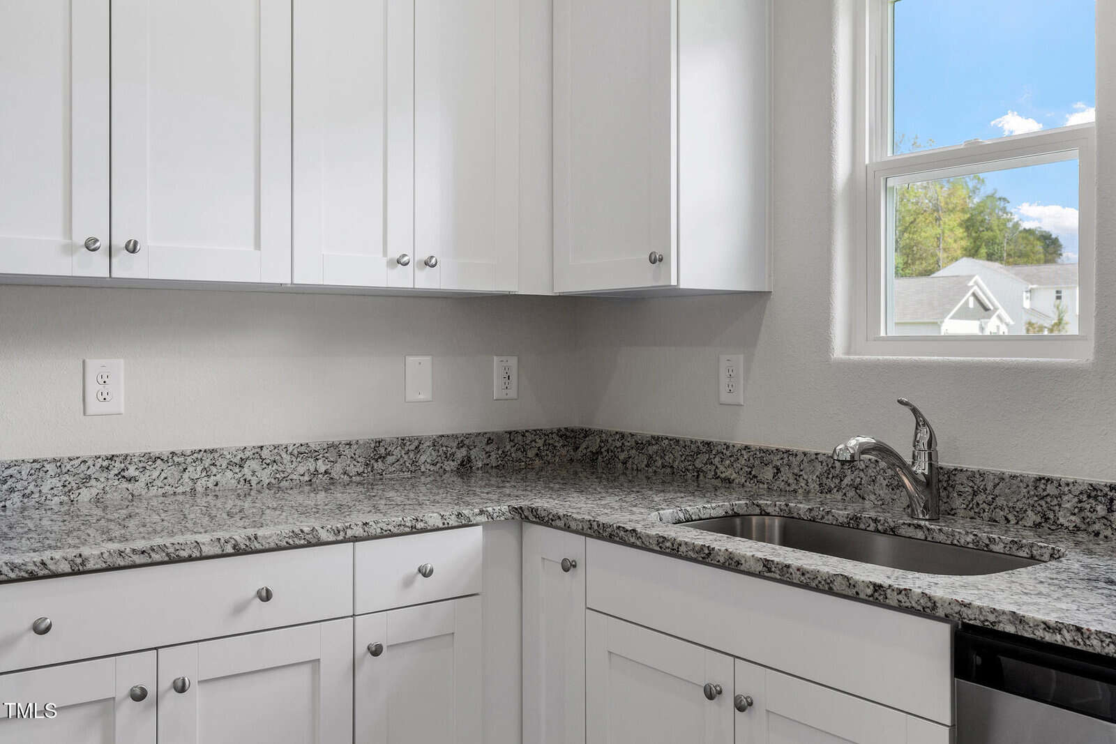 A modern kitchen corner with white cabinets, a speckled granite countertop, and a stainless steel sink. Natural light comes through a window with a view of trees and rooftops. White walls and electrical outlets are visible above the counter.