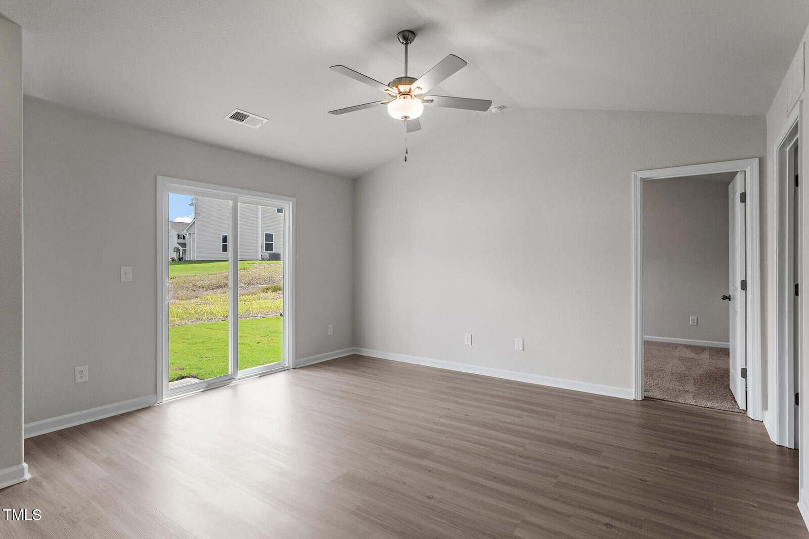 Empty room with light wood flooring, a ceiling fan, and sliding glass doors leading to a grassy backyard. The walls are painted light gray. A carpeted room is visible through an open door on the right.
