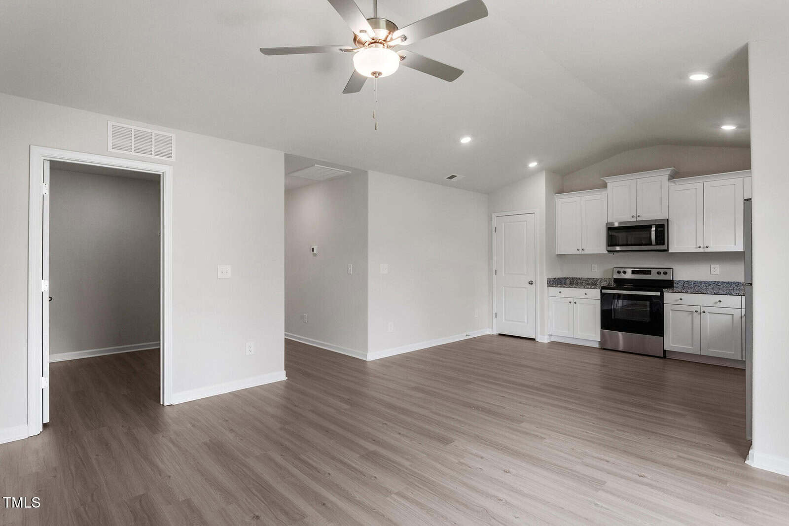 Modern, empty living space with light wood flooring and neutral walls. Features a ceiling fan, recessed lighting, and an open kitchen with stainless steel appliances and white cabinetry. A doorway leads to an adjacent room.