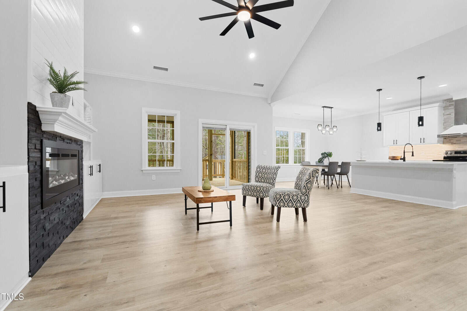 A spacious, modern living area with light wood floors, a black fireplace, two patterned chairs, a wooden coffee table, and an open kitchen with white cabinets, island seating, and pendant lights. Large windows let in natural light.