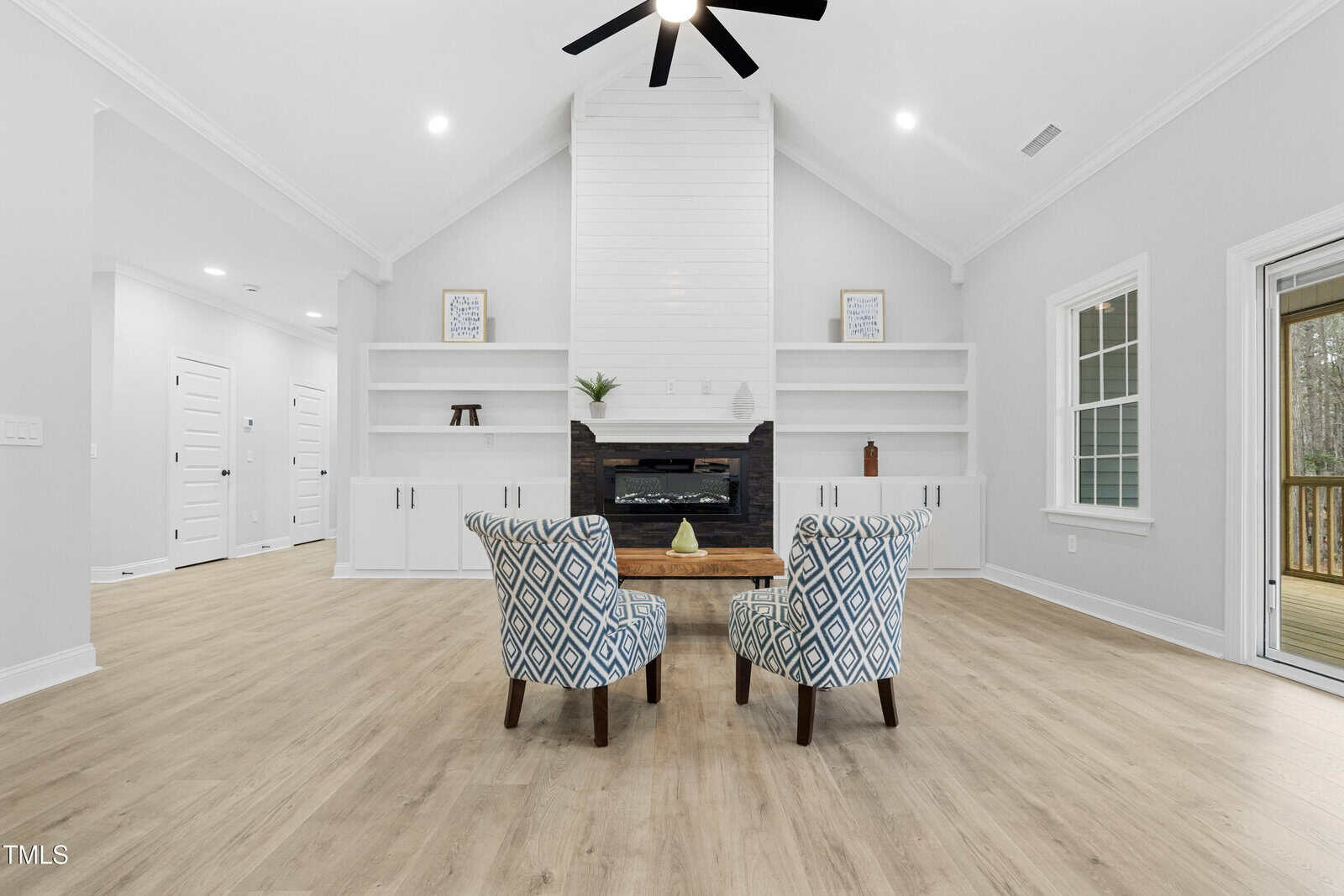 A spacious living room with light wood flooring, white walls, built-in shelves and cabinets, two patterned armchairs facing a wooden table, and a fireplace with a white chimney wall.