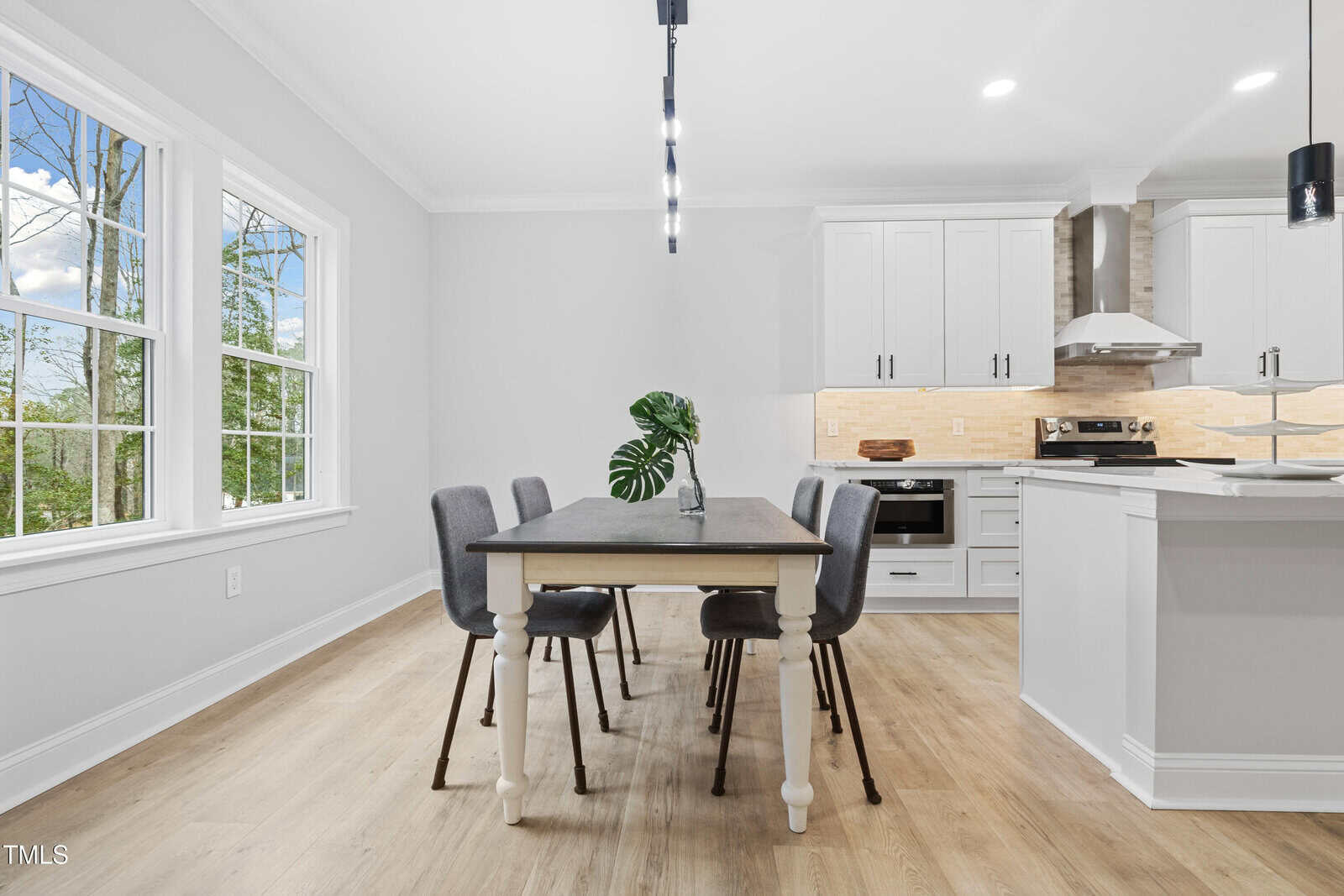 Modern dining area with a rectangular table, four gray chairs, a green plant centerpiece, large windows, and an adjacent white kitchen with stainless steel appliances and wood accents.