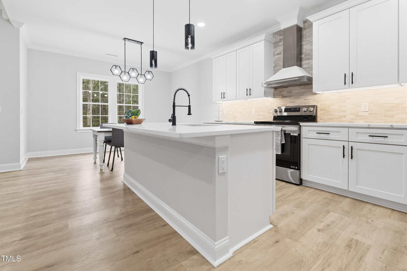 Modern kitchen with white cabinetry, stainless steel appliances, a large island with a black faucet, light wood flooring, pendant lighting, and a dining area with a window in the background.